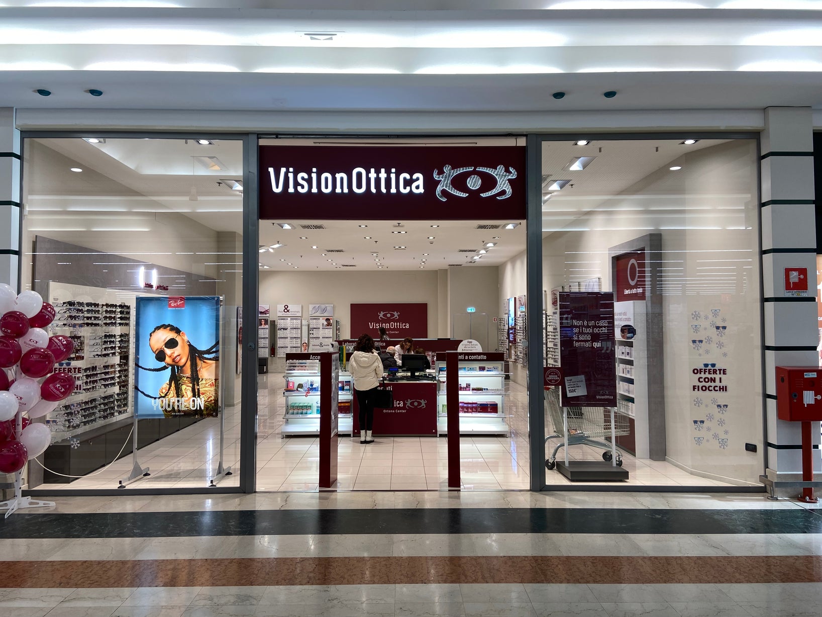 Ingresso del negozio VisionOttica Ortona Center di Ortona in centro commerciale con insegna bordeaux, vetrine laterali con espositori di montature e poster, banner promozionale e personale all'interno