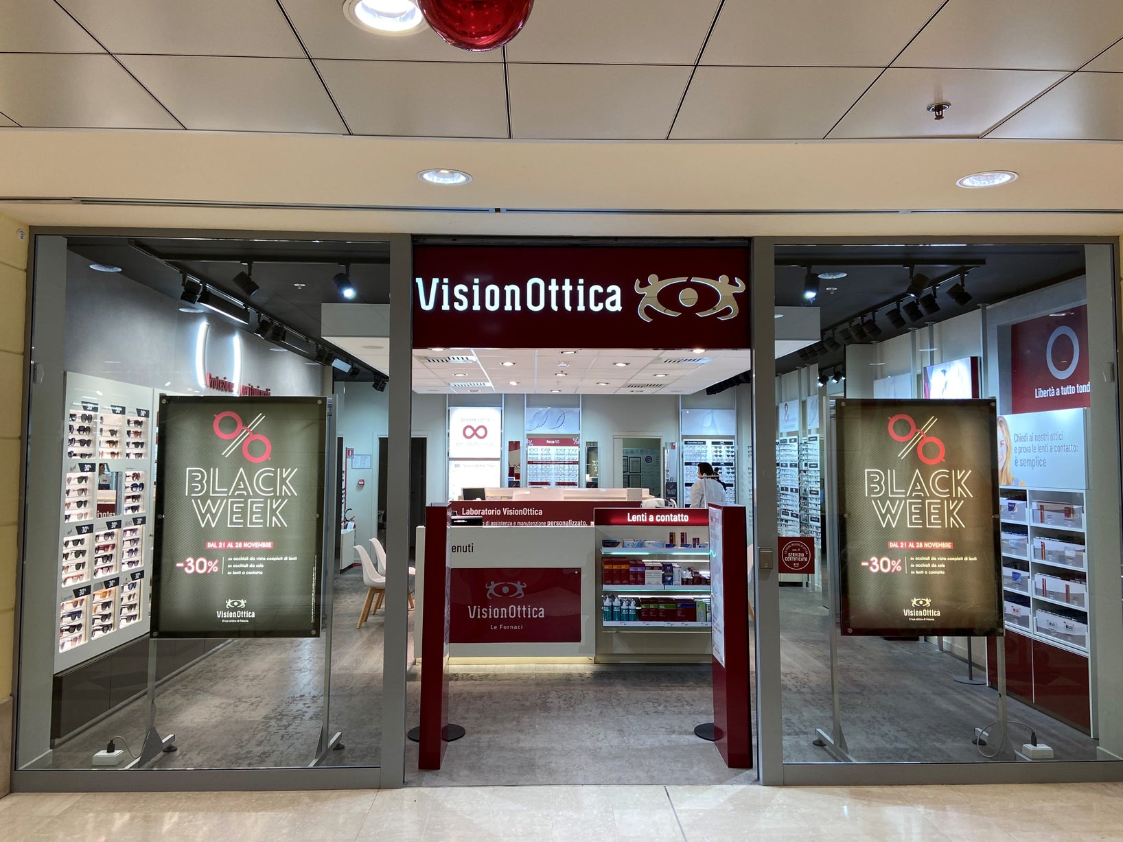 Ingresso del negozio VisionOttica Forlimpopoli di Forlì in centro commerciale con insegna bordeaux, doppi pannelli illuminati Black Week -30% sui lati e bancone con laboratorio ottico visibile