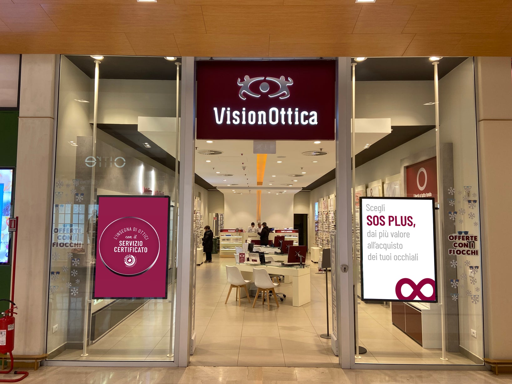 Ingresso del negozio VisionOttica Pescara Nord di Pescara in centro commerciale con insegna bordeaux illuminata su pannello grande, porta a vetri e display promozionali laterali