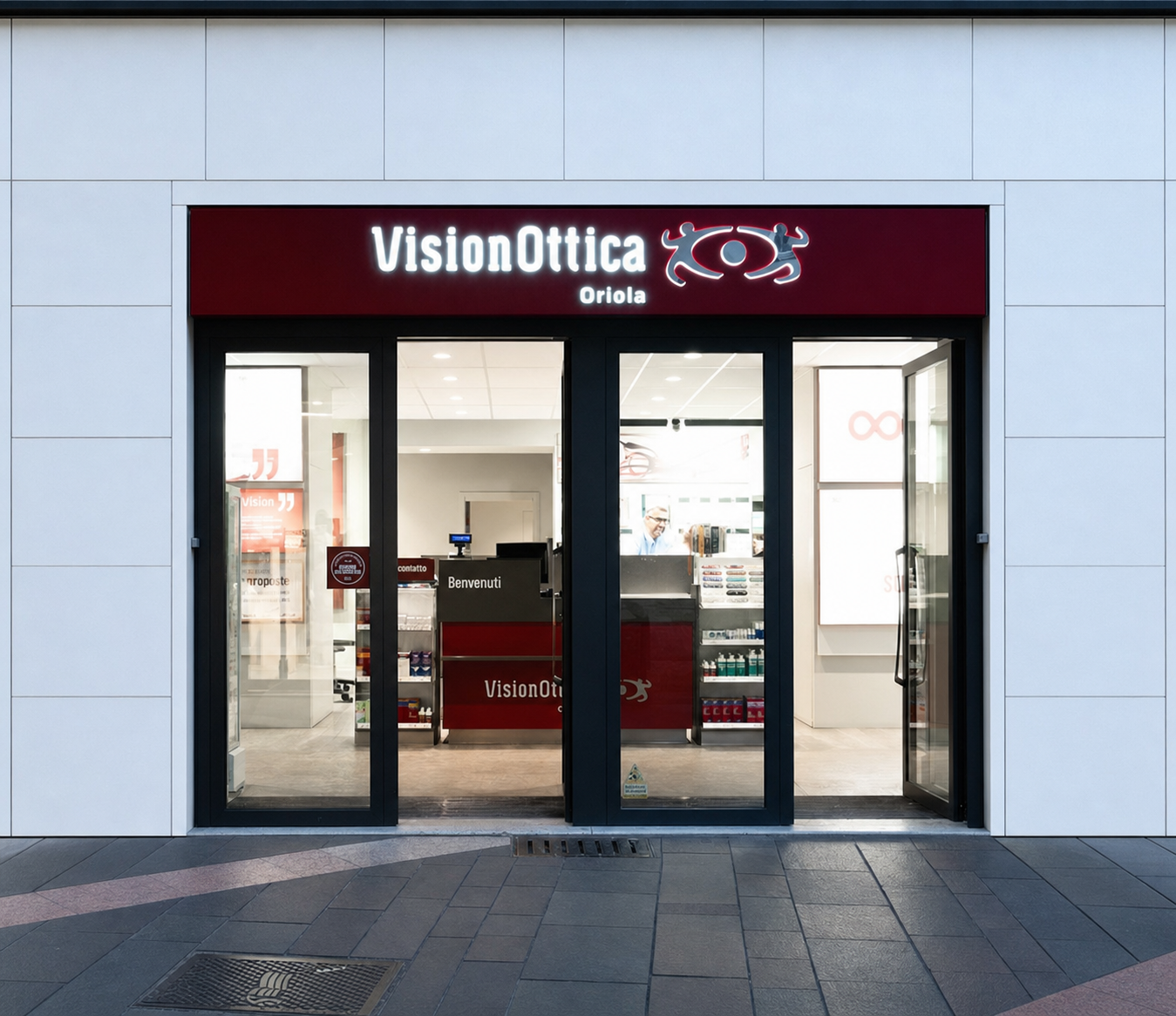 Facciata del negozio VisionOttica Oriola di Trento su strada con insegna bordeaux rettangolare, porta a vetri ampia e interno con bancone centrale visibile