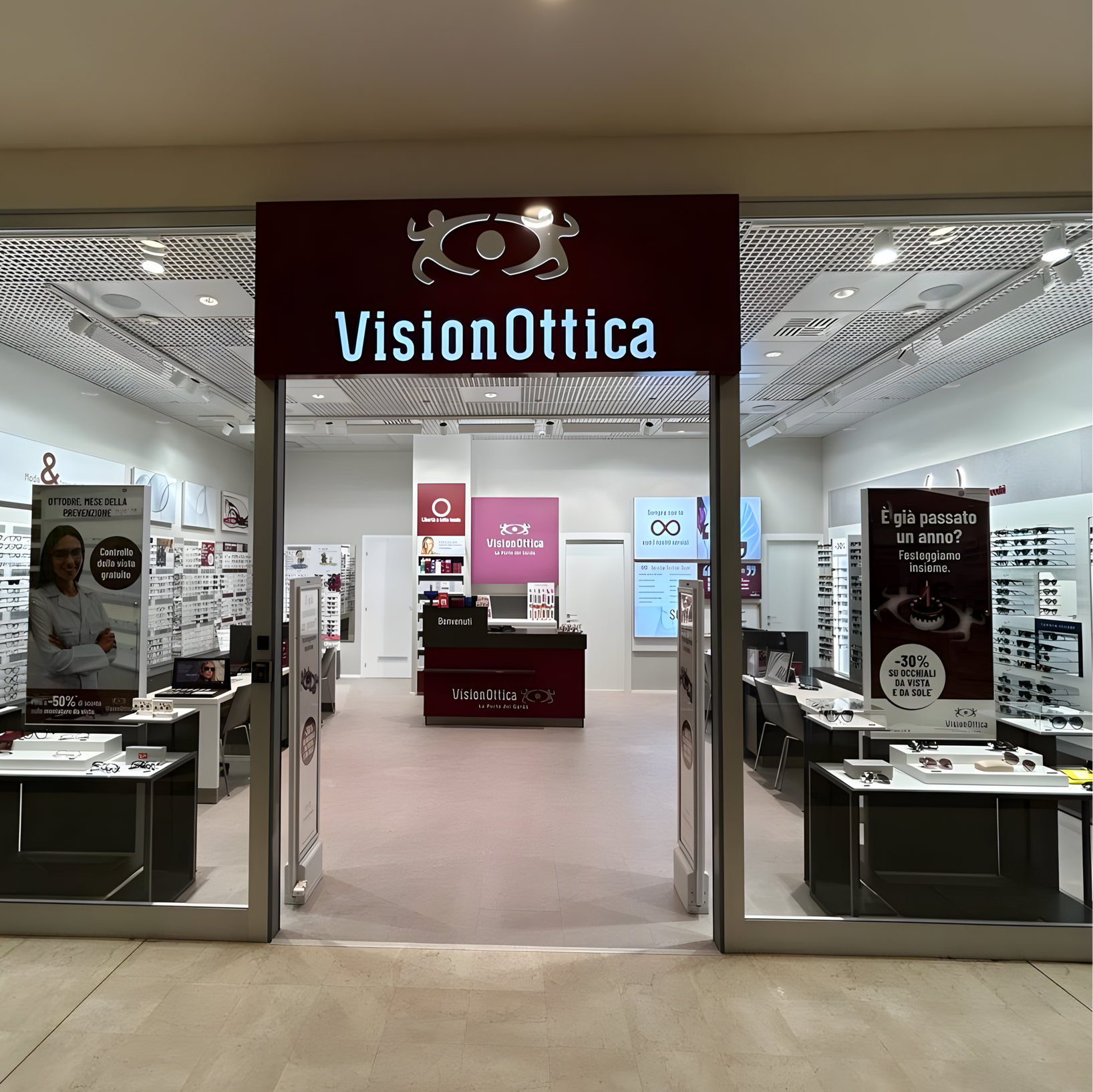 Ingresso del negozio VisionOttica Gavardo Park di Gavardo in centro commerciale con insegna bordeaux con logo luminoso, porta ampia aperta e interno con espositori e totem promozionali laterali