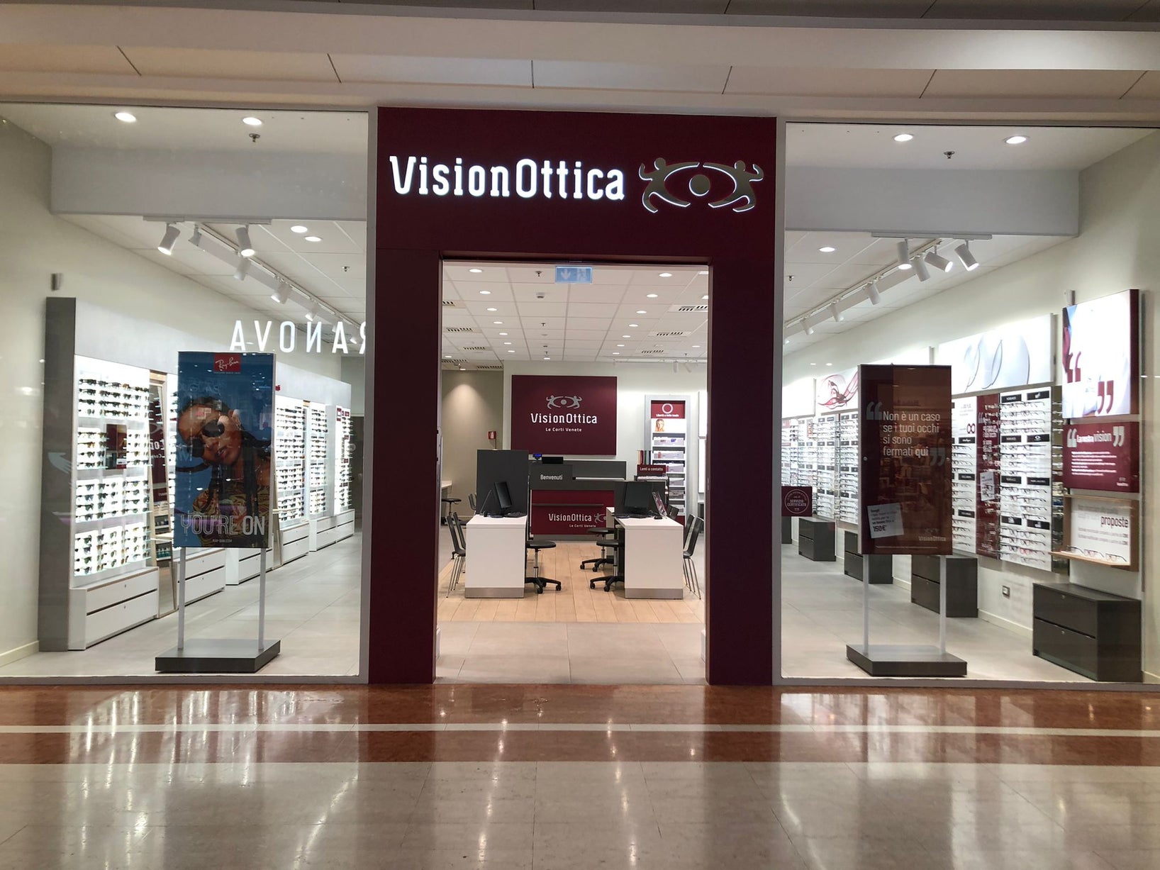 Ingresso del negozio VisionOttica Le Corti Venete di San Martino Buon Albergo in centro commerciale con fascia bordeaux ampia, bancone centrale visibile, espositori di montature su scaffali laterali e totem pubblicitari