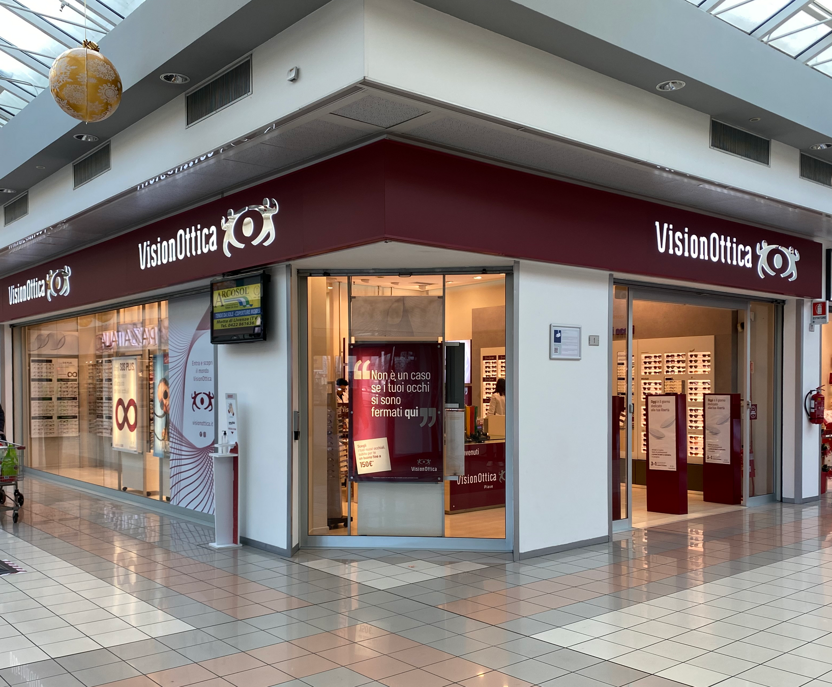 Ingresso del negozio VisionOttica Piave di San Donà di Piave in centro commerciale su angolo con fascia bordeaux continua, due insegne VisionOttica, porta a vetri scorrevole e interno con espositori