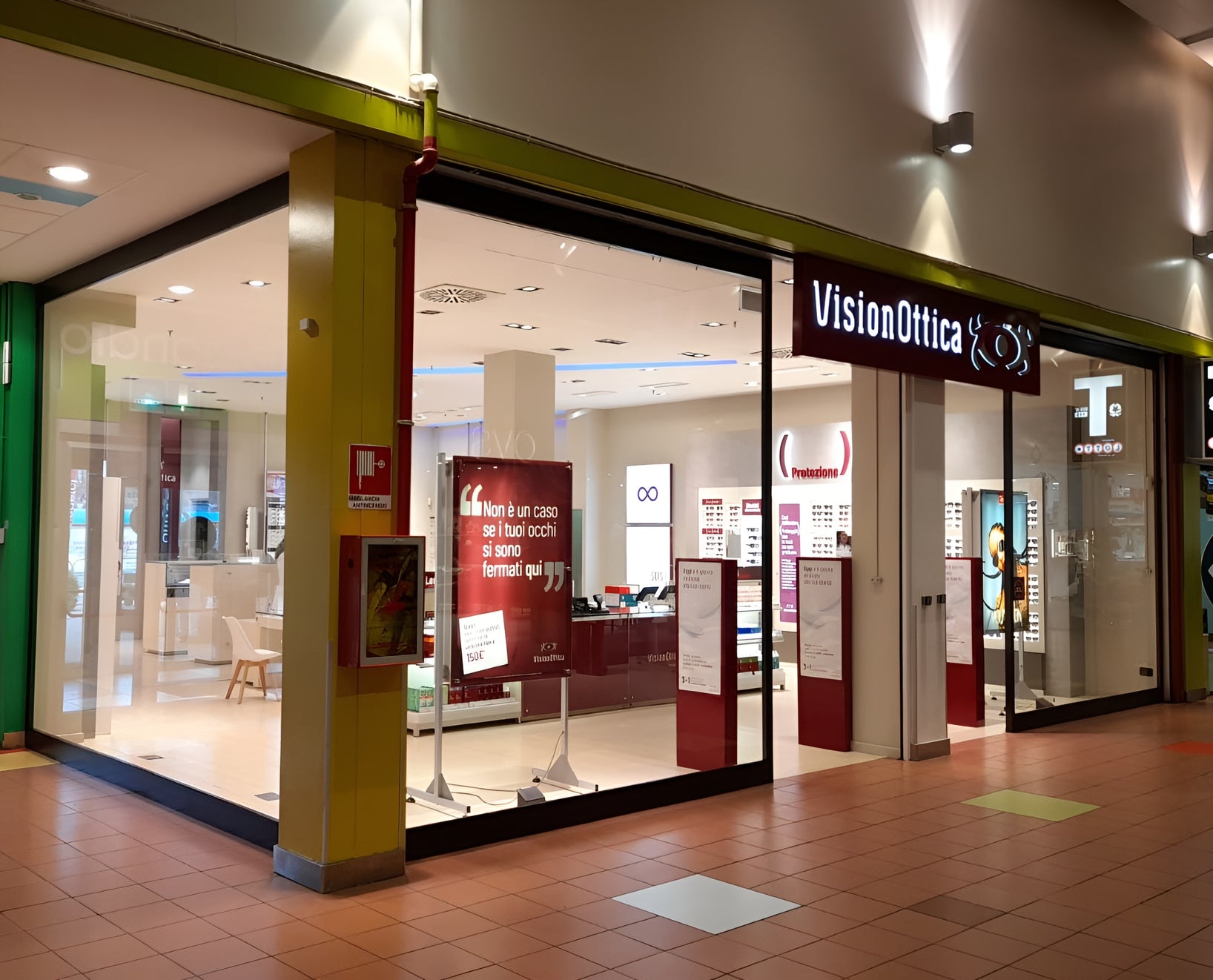 Ingresso del negozio VisionOttica Bergamo di Bergamo in centro commerciale con insegna bordeaux, pilastro verde oliva laterale, vetrina con banner rosso e interno aperto con espositori