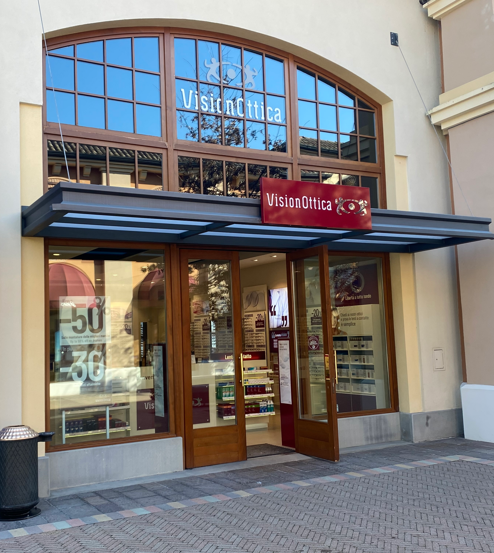Facciata del negozio VisionOttica Fidenza Village di Fidenza in outlet village con architettura ad arco in stile classico, insegna bordeaux, porta in legno a doppia anta aperta e vetrine con sconti esposti