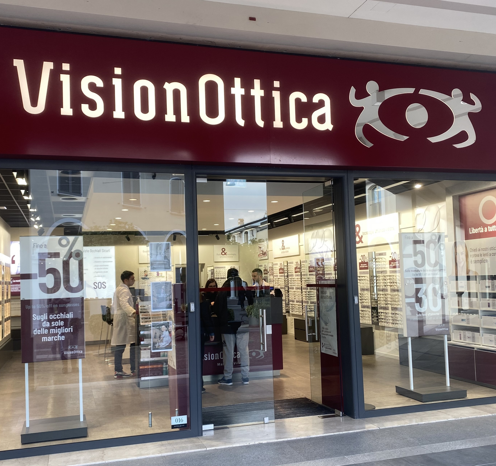 Facciata del negozio VisionOttica Mantova Village di Bagnolo San Vito in outlet village con grande insegna bordeaux, porta scorrevole a vetri, scaffali di occhiali interni visibili e clienti all'ingresso