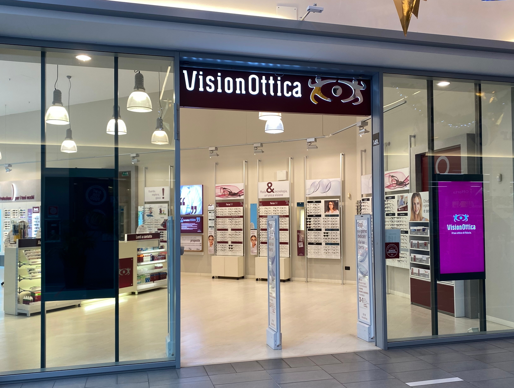 Ingresso del negozio VisionOttica Terminal Nord di Udine in centro commerciale con insegna bordeaux, porta ampia a vetri, interno con espositori di listini ottici a parete e totem digitale VisionOttica
