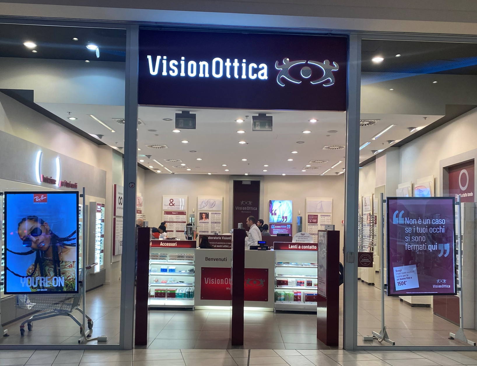 Ingresso del negozio VisionOttica Montedoro di Trieste in centro commerciale con insegna retroilluminata blu, bancone centrale bordeaux e schermi promozionali laterali