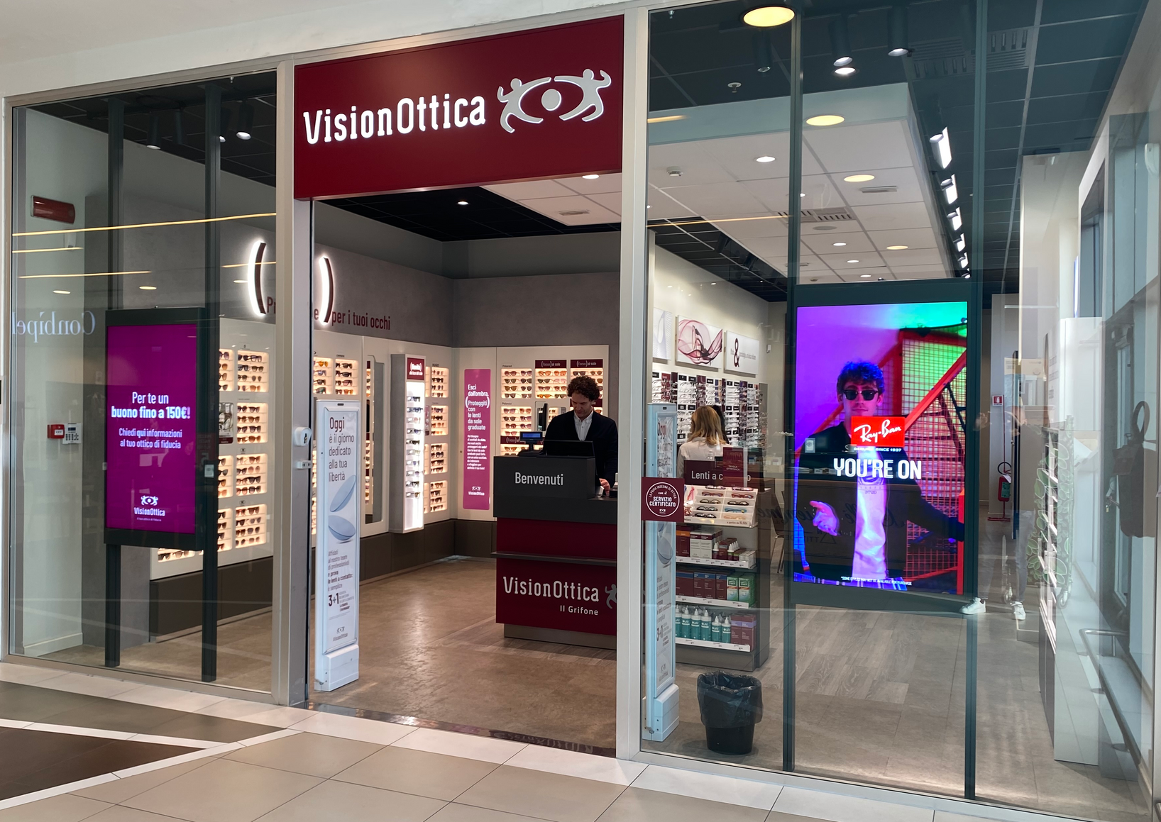 Ingresso del negozio VisionOttica Il Grifone di Bassano del Grappa in centro commerciale con insegna bordeaux, operatori al bancone e schermo Ray-Ban illuminato lateralmente