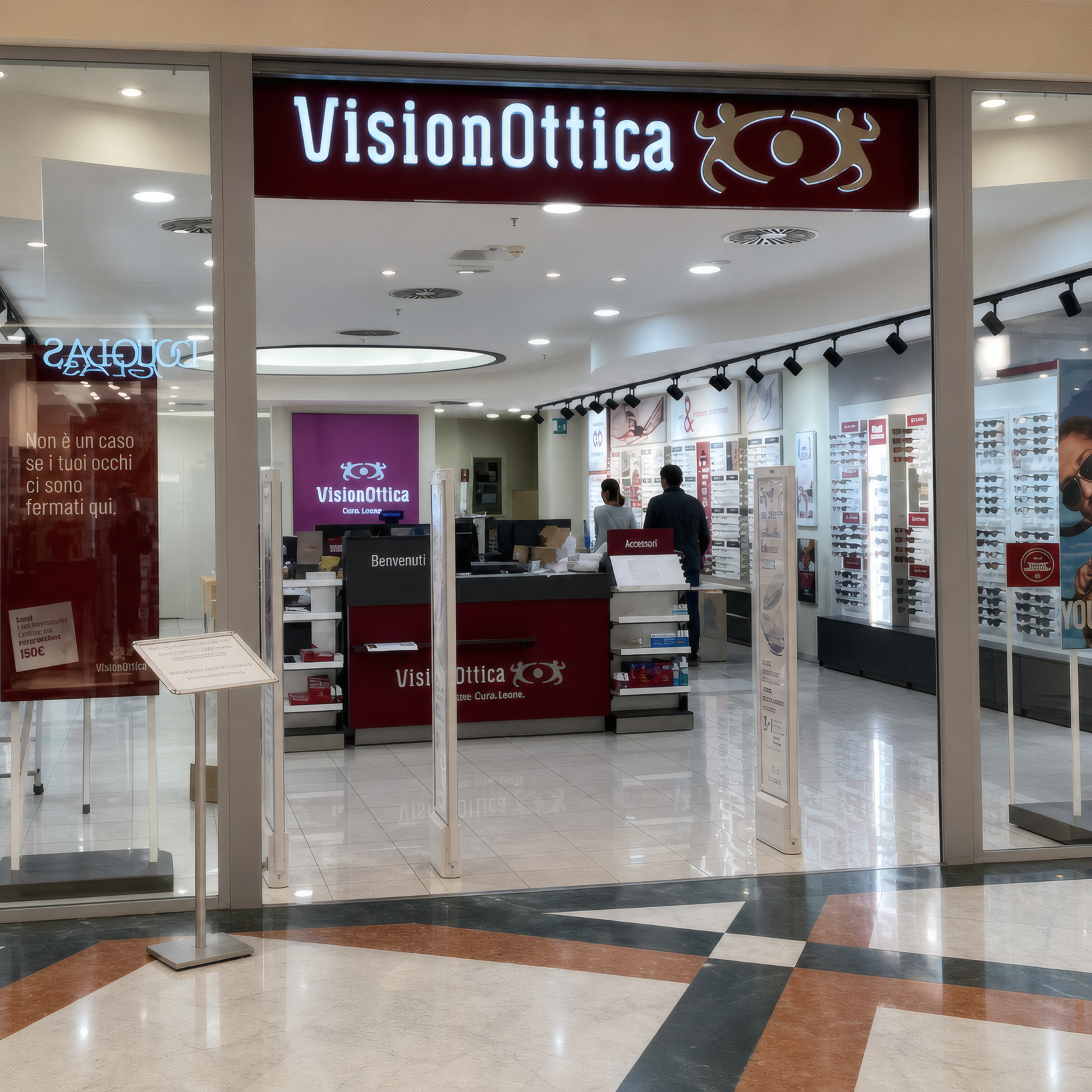 Ingresso del negozio VisionOttica La Corte Lombarda di Bellinzago Lombardo in centro commerciale con pavimento a scacchi, insegna bordeaux e espositori a parete con montature
