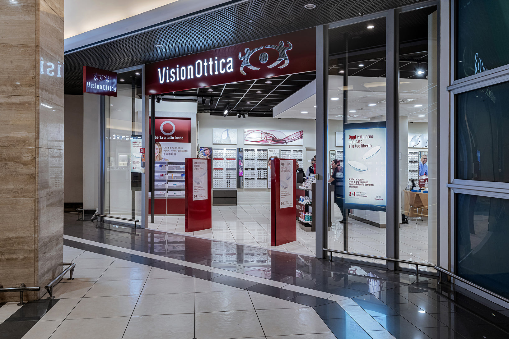 Ingresso del negozio VisionOttica Vulcano di Sesto San Giovanni in centro commerciale con ampia vetrata, totem promozionali rossi e insegna bordeaux su soffitto scuro