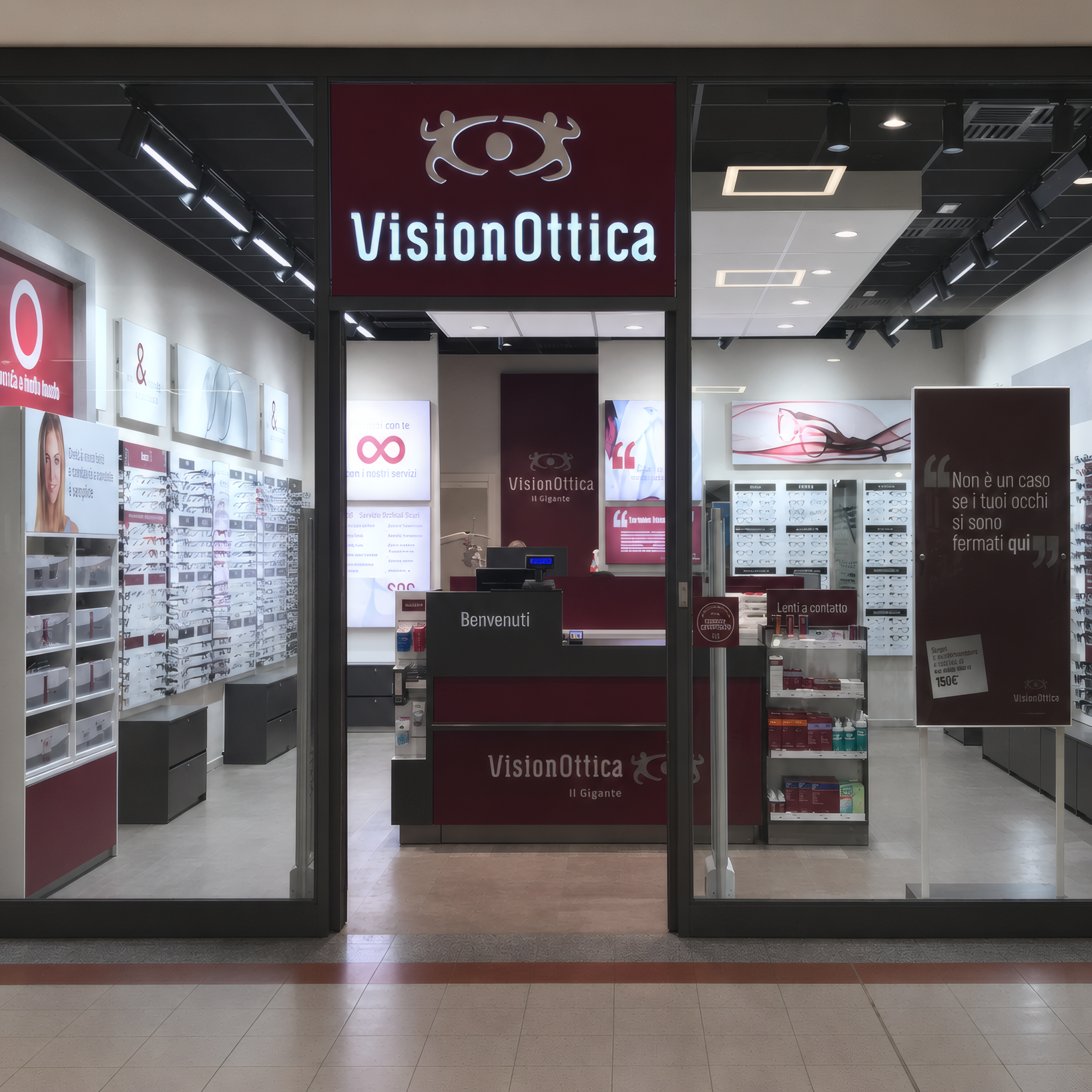 Ingresso del negozio VisionOttica Villasanta in centro commerciale con insegna bordeaux su fondo scuro, vetrine laterali con espositori a parete e bancone centrale