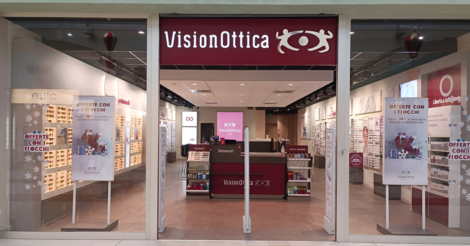 Vetrina del negozio VisionOttica Pieve di Pieve Fissiraga in centro commerciale con insegna bordeaux, pannelli natalizi Offerte con Fiocchi e espositori di occhiali visibili attraverso il vetro