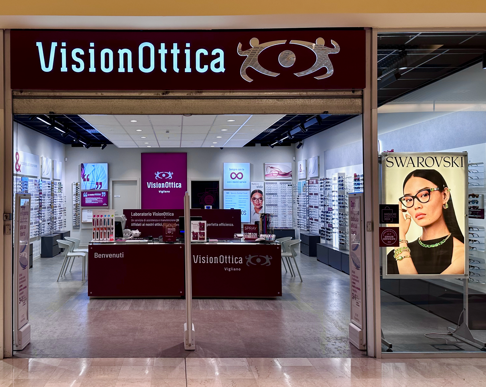 Ingresso del negozio VisionOttica Vigliano di Vigliano Biellese in centro commerciale con insegna luminosa azzurra, parete espositiva con montature e poster Swarovski laterale
