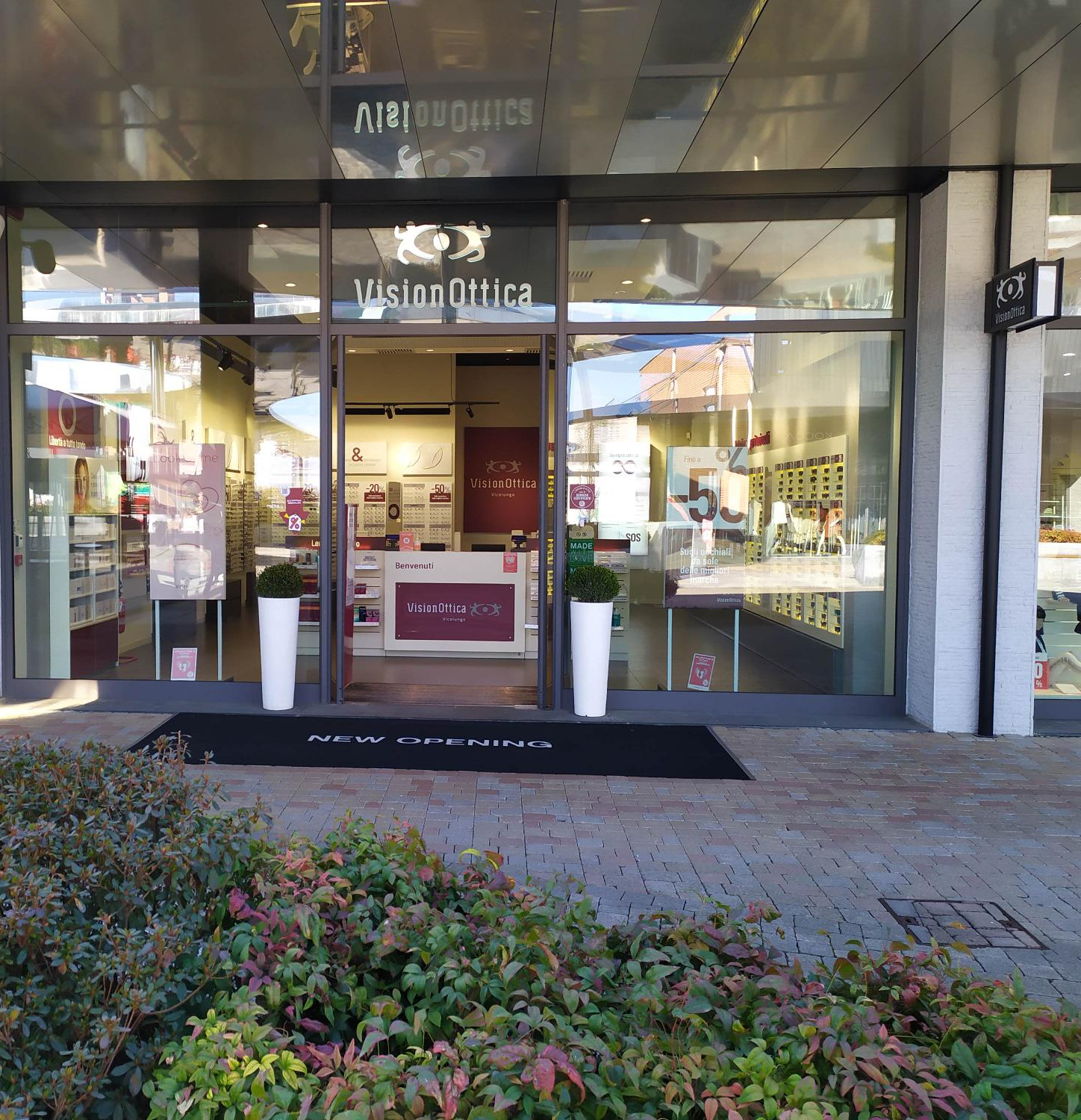 Facciata del negozio VisionOttica Vicolungo su strada commerciale a Vicolungo con ingresso a vetri su piazzale pavimentato, vasi con piante all'ingresso e tappeto New Opening