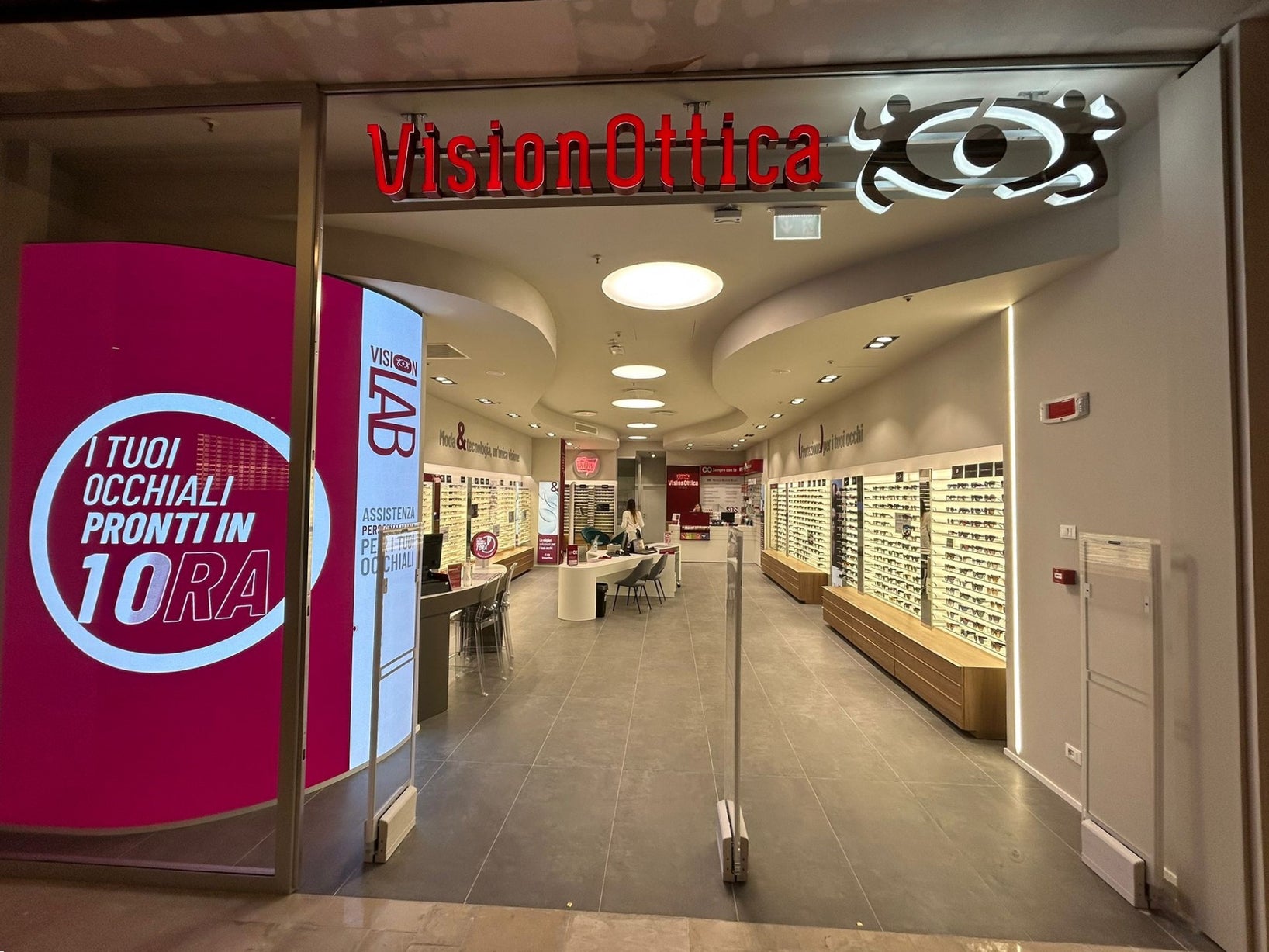 Ingresso del negozio VisionOttica Le Gru di Grugliasco in centro commerciale con insegna rossa retroilluminata, schermo totem con promozione Pronti in 10 Ore e ampi espositori interni