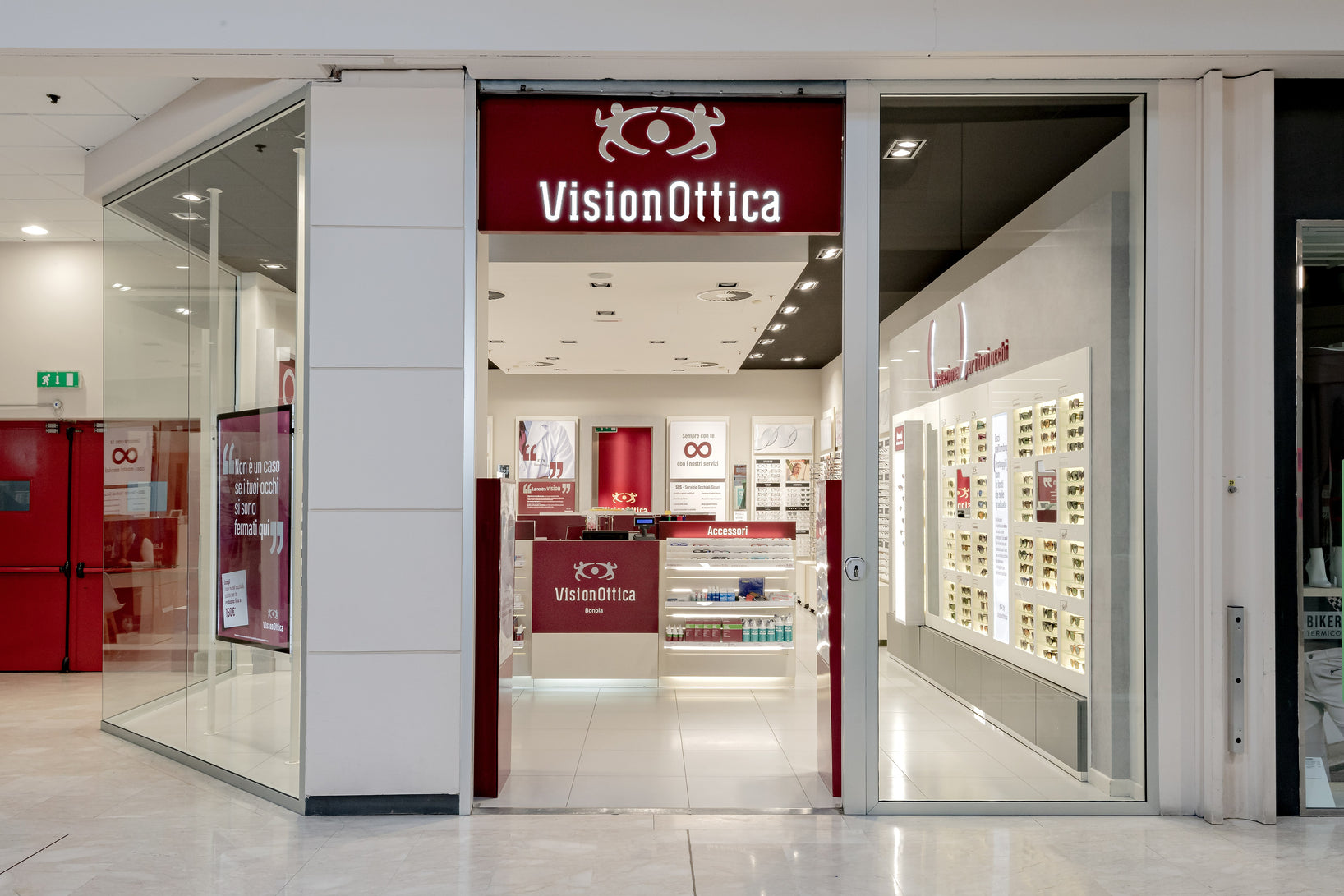 Ingresso del negozio VisionOttica Bonola di Milano in centro commerciale con insegna bordeaux, porta a vetri centrale e parete espositiva con montature da vista visibile internamente