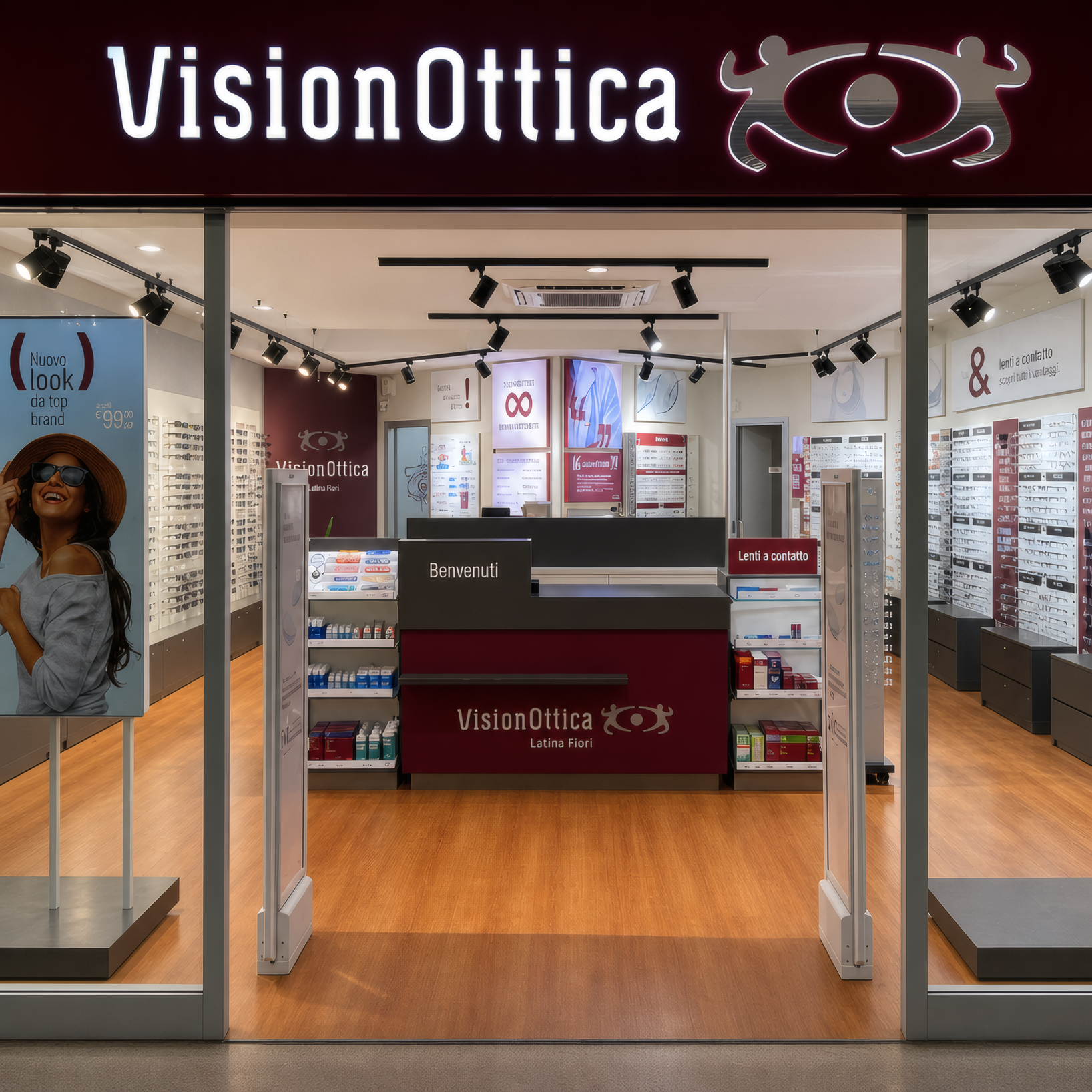 Ingresso del negozio VisionOttica Latinafiori di Latina in centro commerciale con insegna bordeaux, bancone centrale e espositori a parete con montature su pavimento in parquet