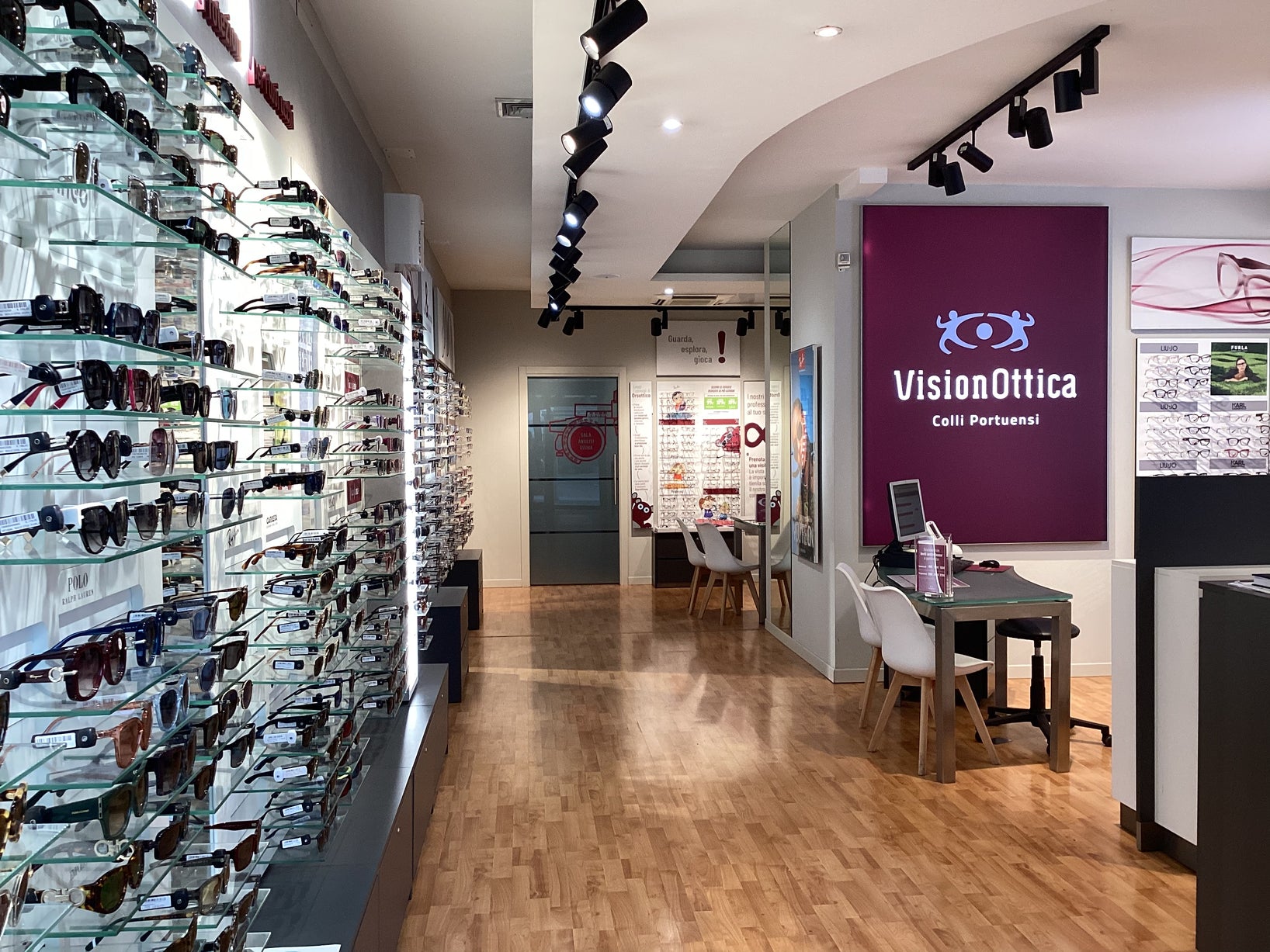 Interno del negozio VisionOttica Colli Portuensi di Roma su strada con parete espositiva a tutta altezza con occhiali da sole, tavolo consulenza e insegna bordeaux a parete