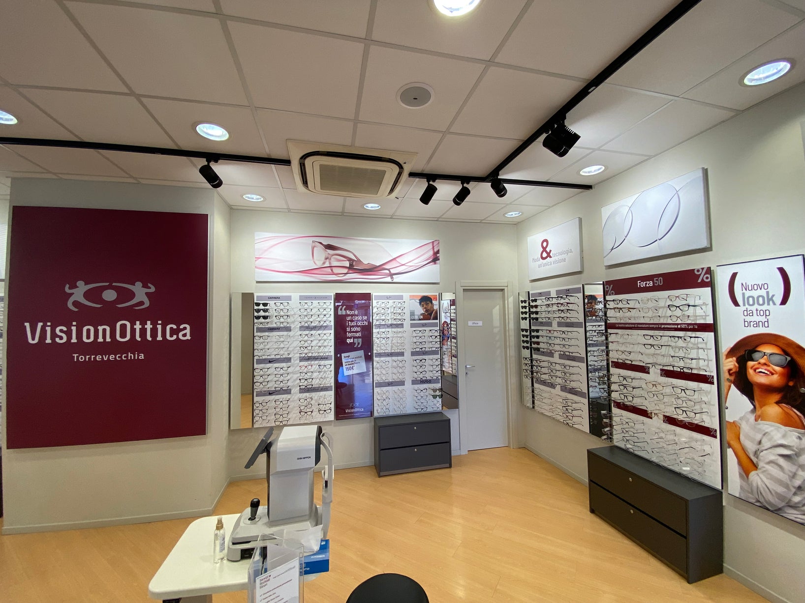 Interno del negozio VisionOttica Torrevecchia di Roma su strada con pareti espositive di montature, pannello promozionale Forza 50% e insegna bordeaux con logo a parete
