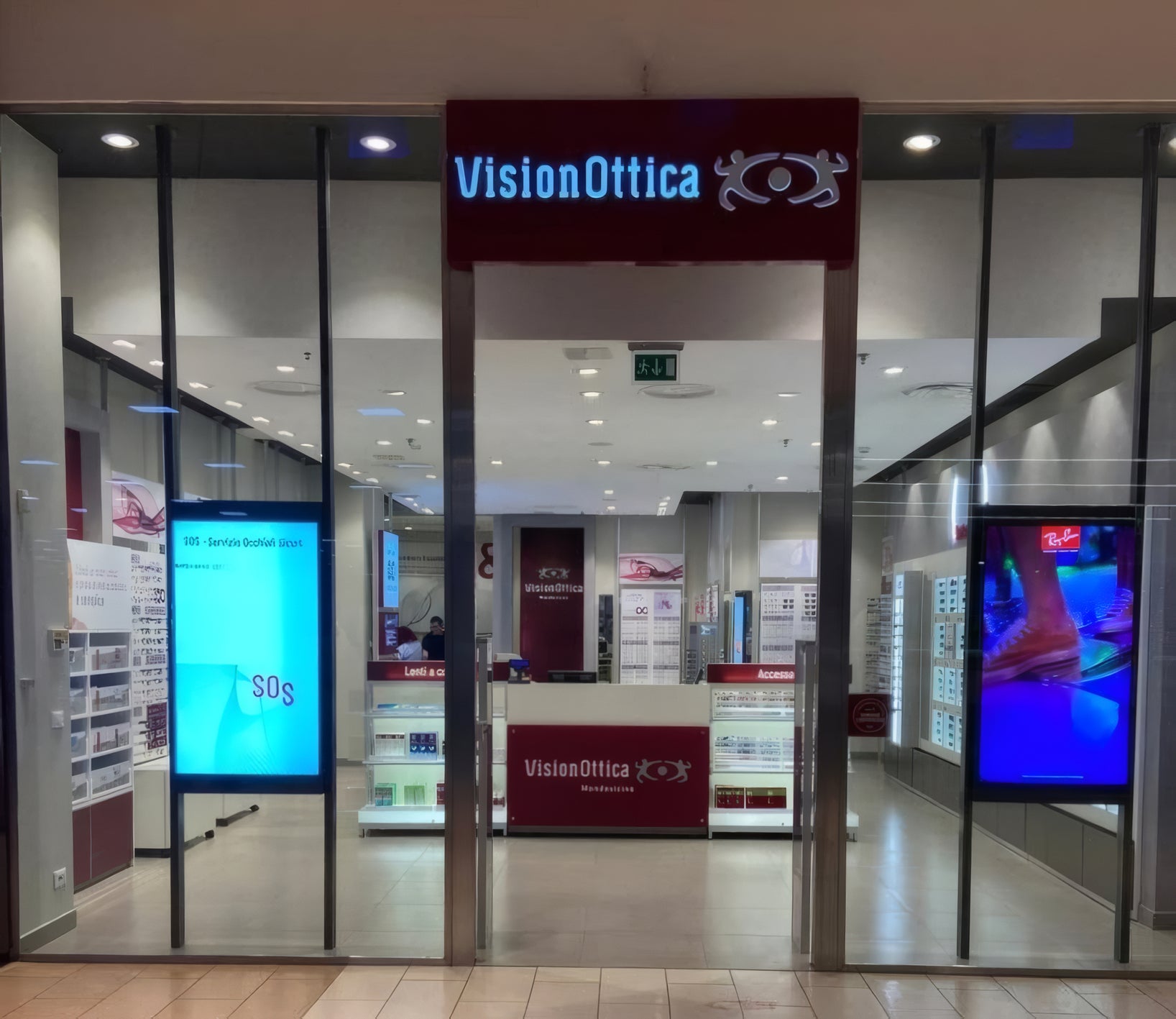 Ingresso del negozio VisionOttica Mondovicino di Mondovì in centro commerciale con insegna bordeaux retroilluminata, vetrate laterali e schermi digitali interni con SOS e Ray-Ban