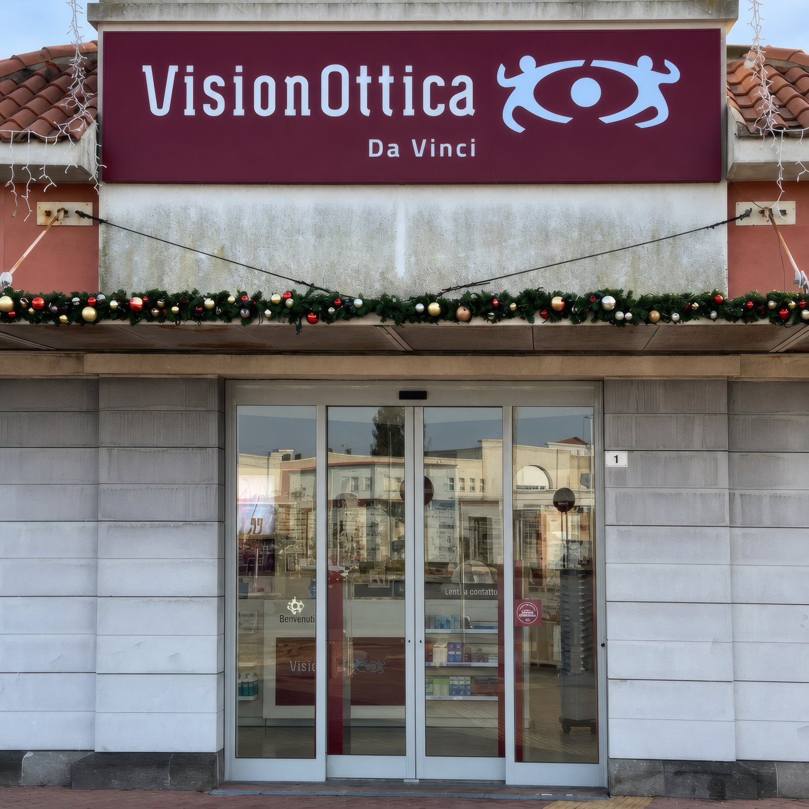 Facciata del negozio VisionOttica Da Vinci di Fiumicino su strada con insegna bordeaux, porta a vetri centrale automatica e decorazioni natalizie con ghirlande sull'architrave