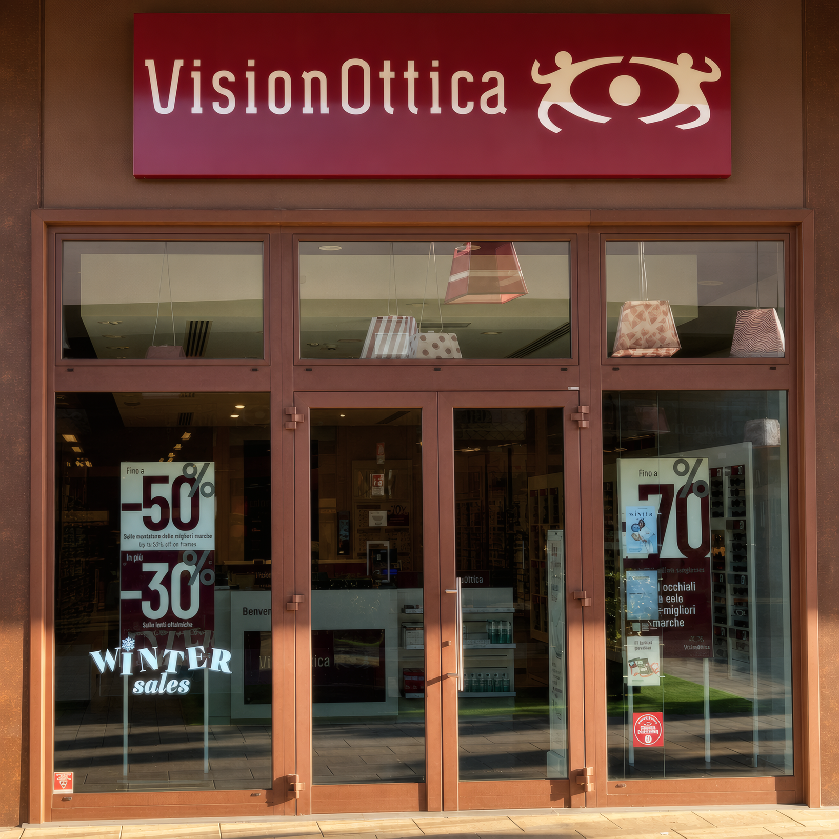 Facciata del negozio VisionOttica 5 Terre di Brugnato in outlet village con porta a vetri su telaio bronzo, insegna bordeaux e vetrina con saldi Winter Sales fino al 70%