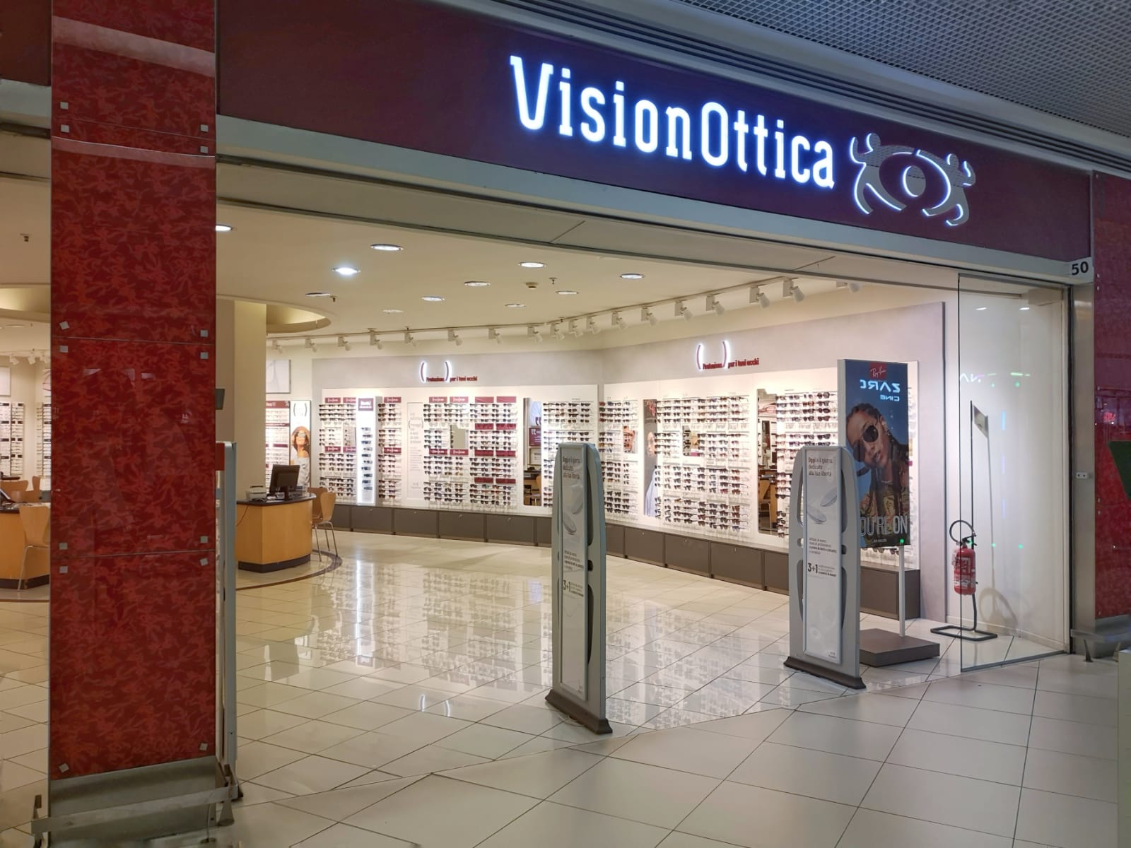 Ingresso del negozio VisionOttica Le Porte di Napoli di Afragola in centro commerciale con insegna luminosa bordeaux, ampie vetrate aperte su espositori di occhiali su pareti bianche e pilastri rossi