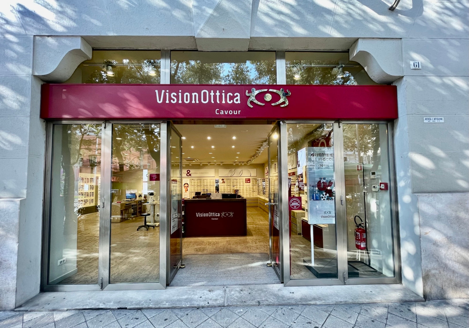Facciata del negozio VisionOttica Cavour di Bari su via pedonale con insegna bordeaux, porta a vetri scorrevole aperta e vetrina che mostra espositori di occhiali e bancone interno illuminato