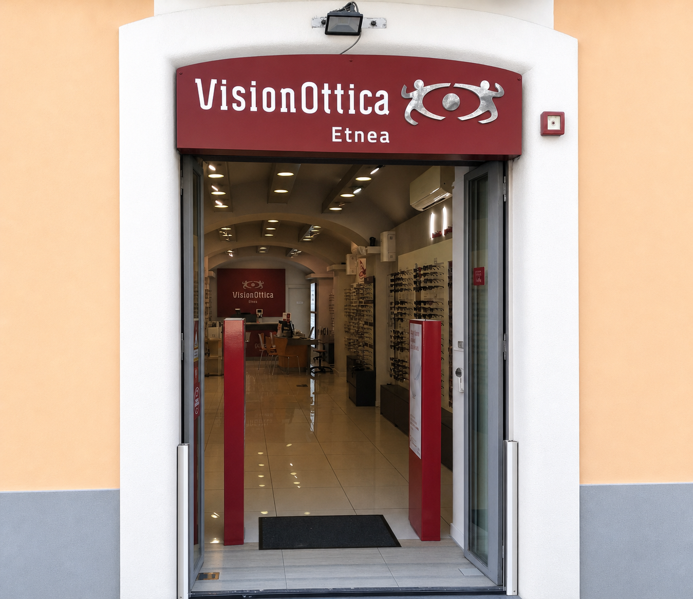 Ingresso del negozio VisionOttica Etnea di Catania in centro storico con insegna bordeaux su edificio intonacato, porta a vetri aperta su interno con espositori di montature e pannello rosso VisionOttica