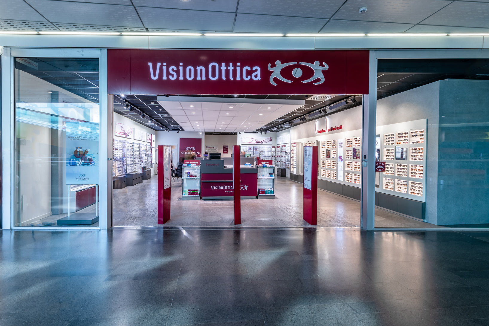 Ingresso del negozio VisionOttica Etnapolis di Belpasso in centro commerciale con insegna bordeaux, ampia apertura aperta su interno moderno con bancone rosso centrale, espositori a parete di occhiali e pavimento scuro lucido