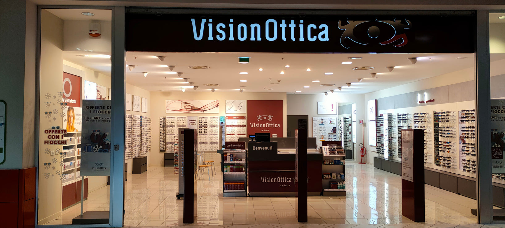 Ingresso del negozio VisionOttica La Torre di Palermo in centro commerciale con insegna luminosa su fondo scuro, interno aperto con lunghi espositori laterali di occhiali, bancone bordeaux centrale con scritta Benvenuti e banner promozionali