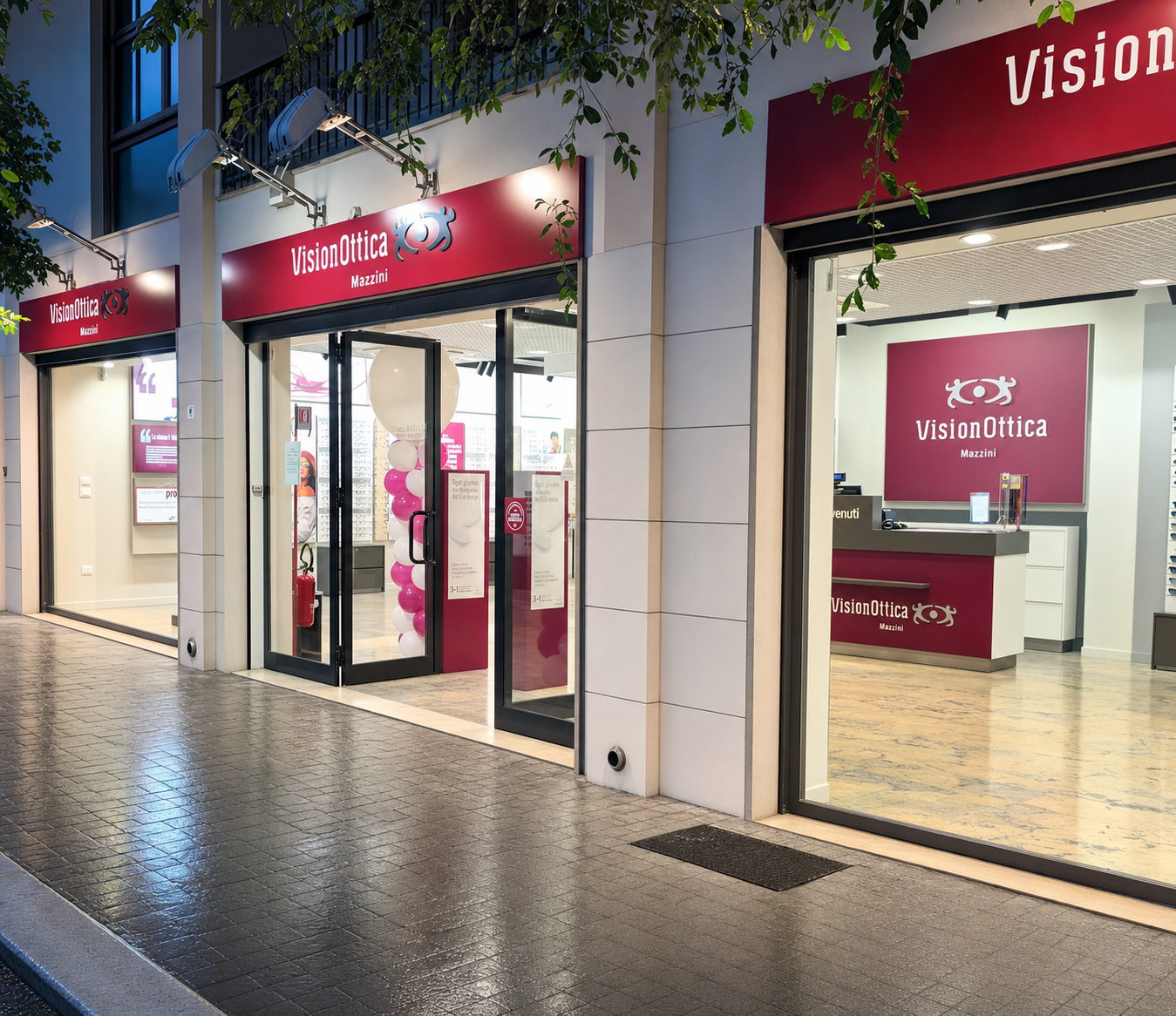 Facciata notturna del negozio VisionOttica Mazzini di Marsala su via pedonale con insegna bordeaux illuminata, porta a vetri chiusa affiancata da ampie vetrine con decorazioni a palloncini rosa e bianchi, interno con bancone visibile