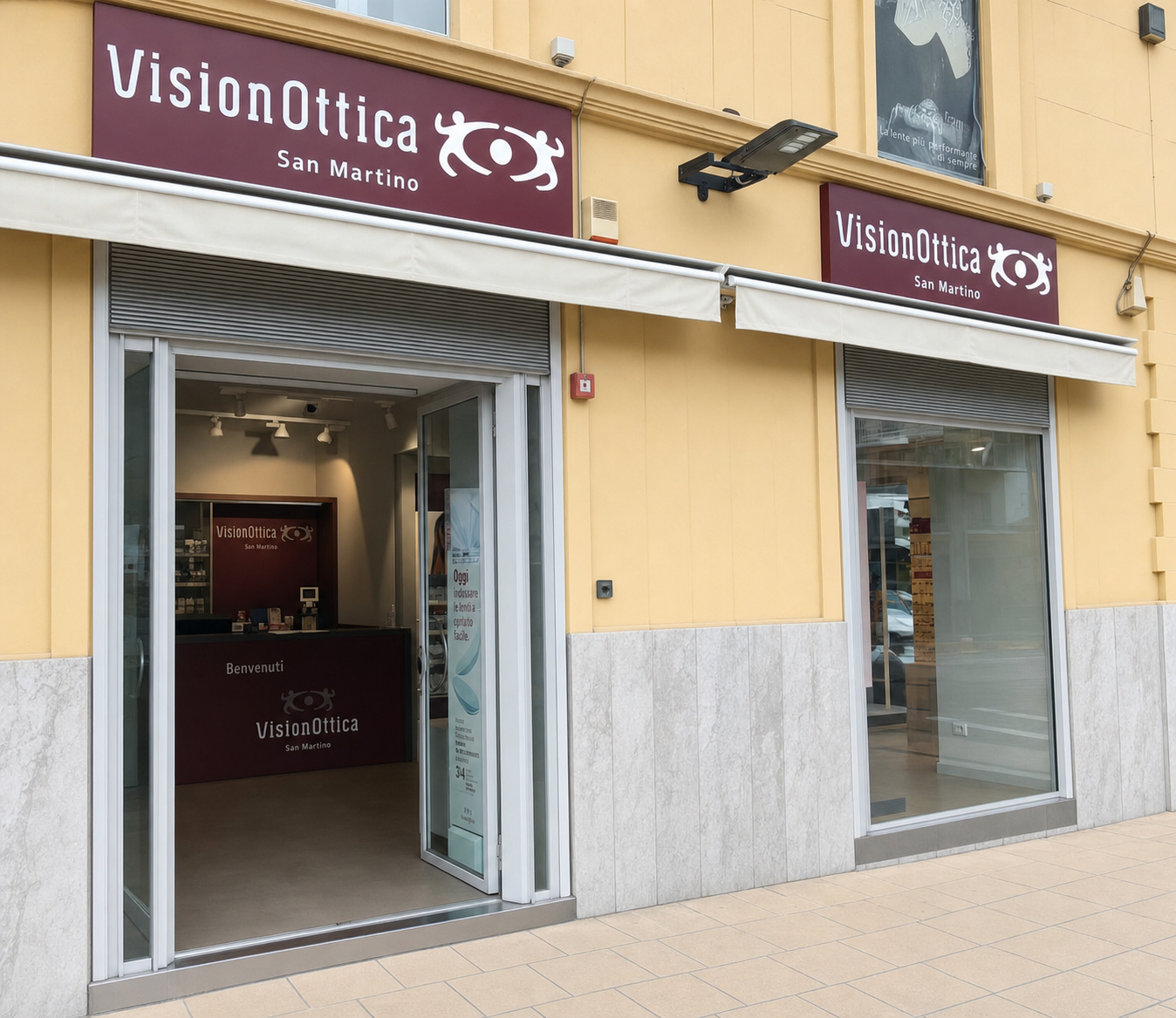 Facciata del negozio VisionOttica San Martino di Messina su strada con due insegne bordeaux affiancate, due ingressi a vetri separati da pilastro su edificio intonacato giallo, interno con bancone bordeaux e scritta Benvenuti visibile