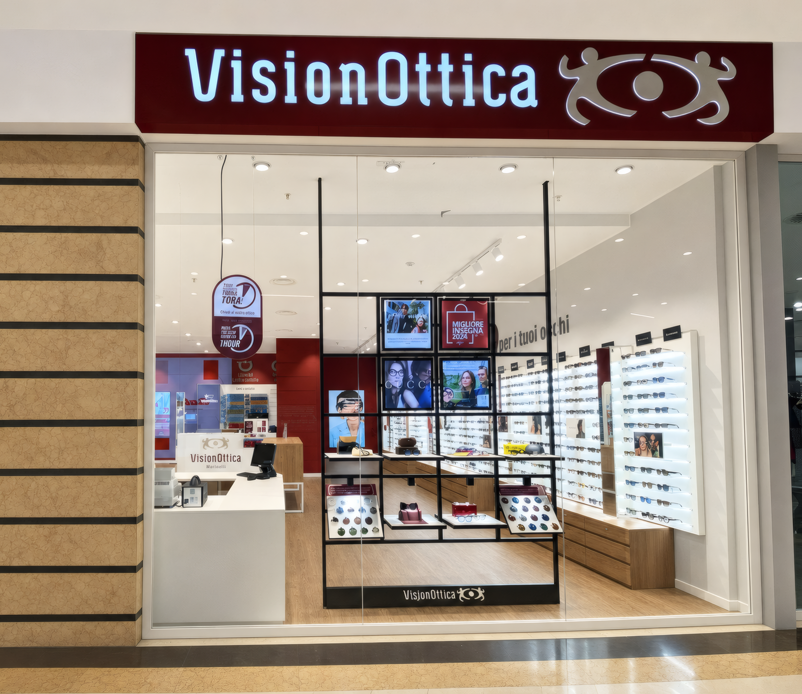 Vetrina del negozio VisionOttica Maximall di Pontecagnano in centro commerciale con insegna bordeaux luminosa, espositore centrale a griglia metallica con schermi digitali e montature esposte, pareti laterali con scaffali di occhiali
