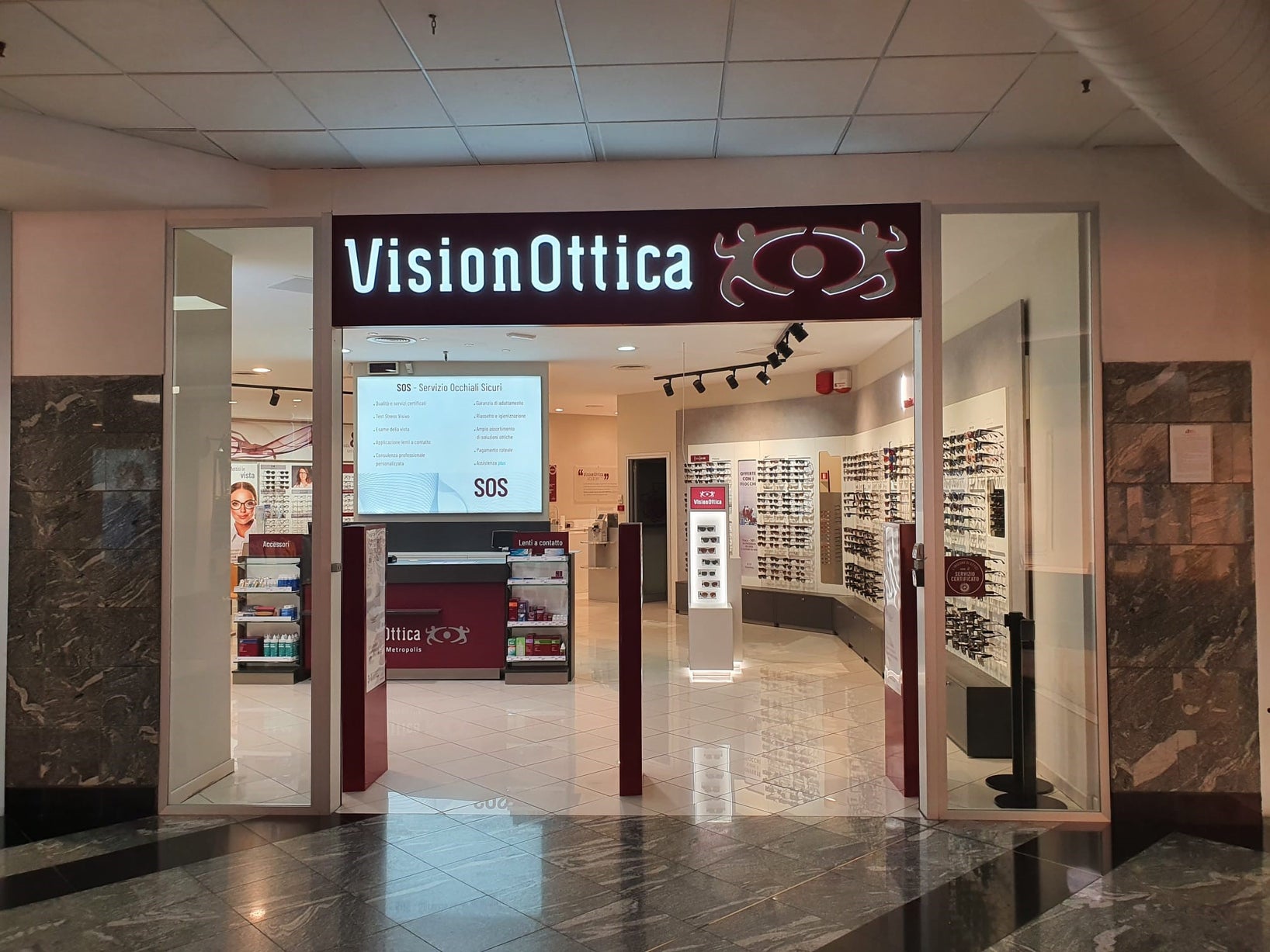 Ingresso del negozio VisionOttica Metropolis di Rende in centro commerciale con insegna bordeaux luminosa, apertura ampia su interno con espositori laterali di occhiali, bancone centrale bordeaux, schermo con presentazione SOS Occhiali Sicuri