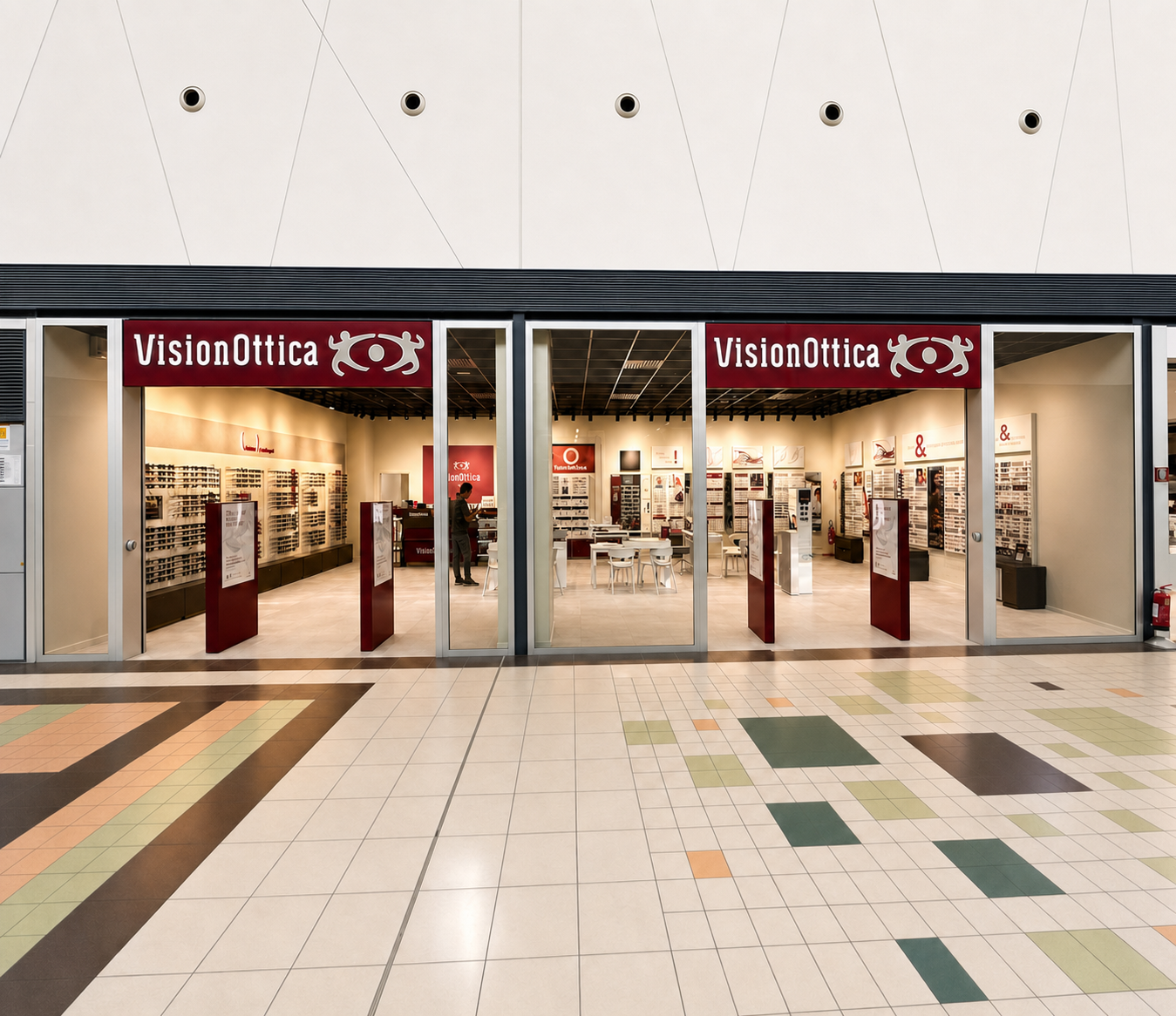 Facciata del negozio VisionOttica Bariblu di Triggiano in centro commerciale con due ampie vetrate affiancate e doppia insegna bordeaux, interno spazioso con lunghi scaffali di montature su entrambi i lati e pavimento a riquadri colorati