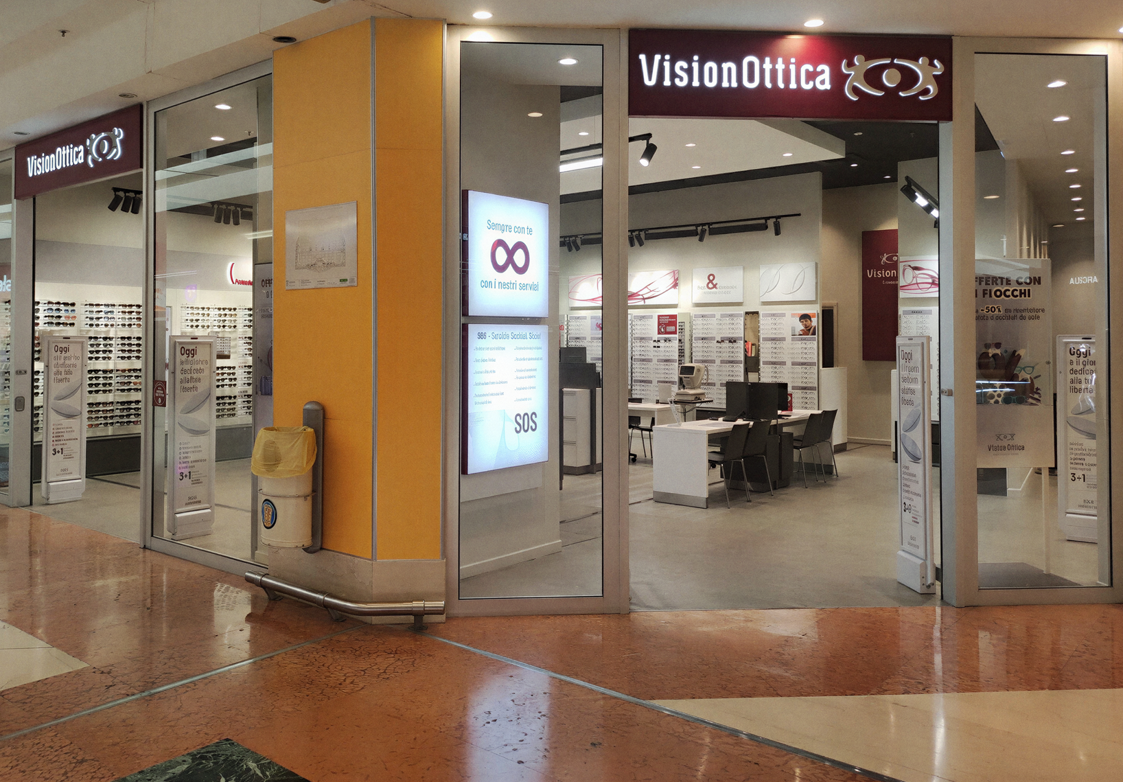 Ingresso del negozio VisionOttica Grande Sud di Giuliano in centro commerciale con insegna bordeaux su pilastro arancione, due aperture separate con espositori di montature e totem luminoso SOS visibile nell'interno
