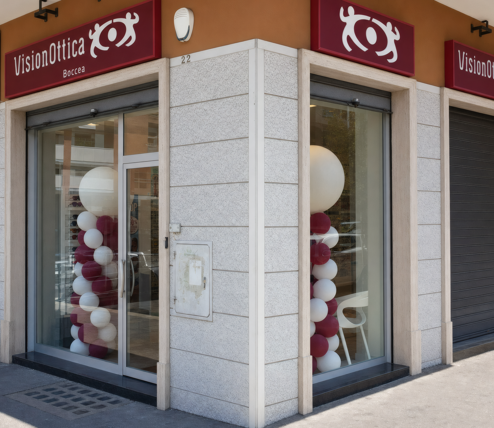 Facciata angolare del negozio VisionOttica Boccea di Roma su strada con due insegne bordeaux e porta a vetri incernierata decorata con colonne di palloncini bordeaux e bianchi su rivestimento in granito grigio chiaro