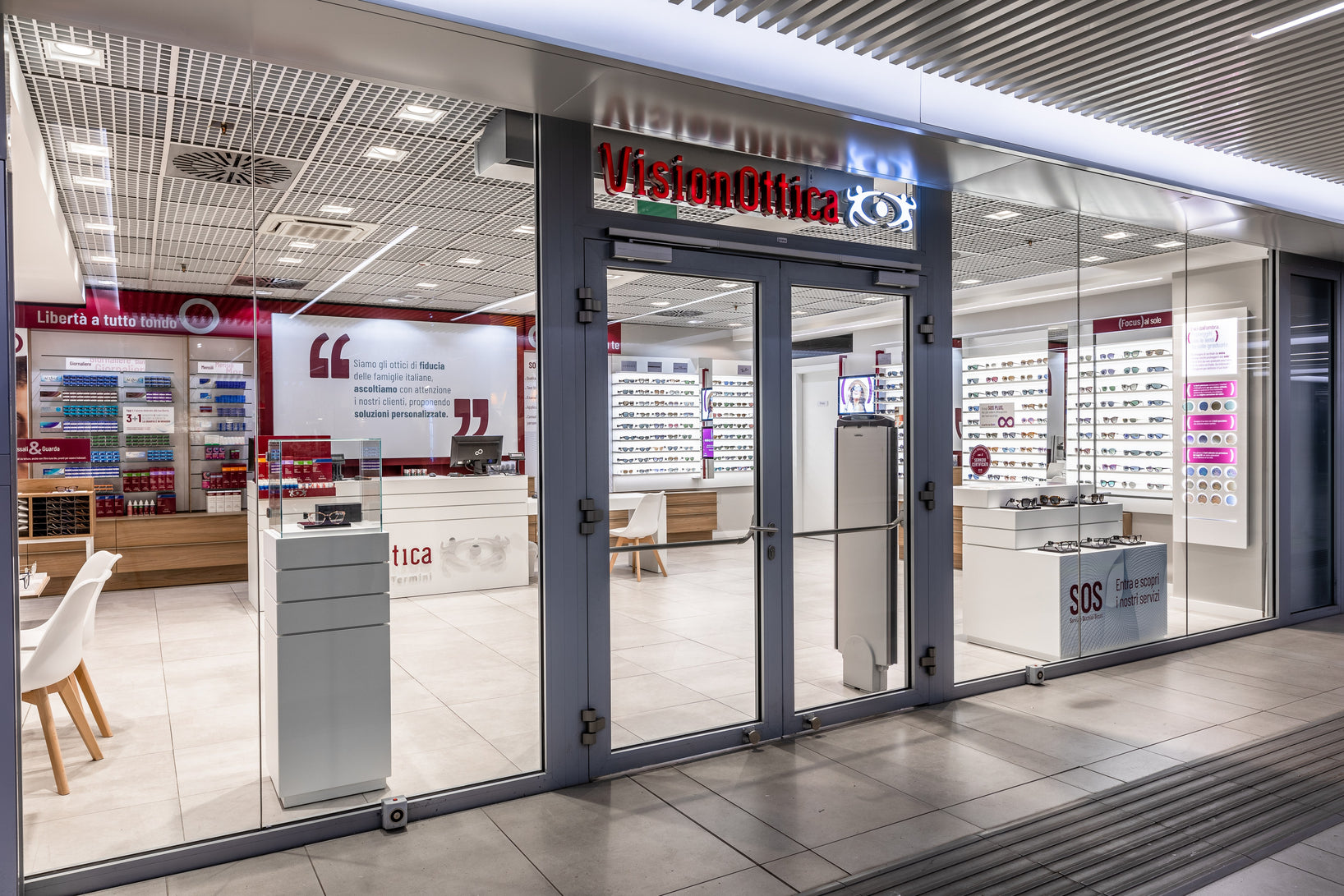 Ingresso del negozio VisionOttica Termini di Roma in galleria con insegna bordeaux retroilluminata, porta a vetri scorrevole affiancata da espositori di occhiali da vista, totem SOS e parete interna con citazione in rosso