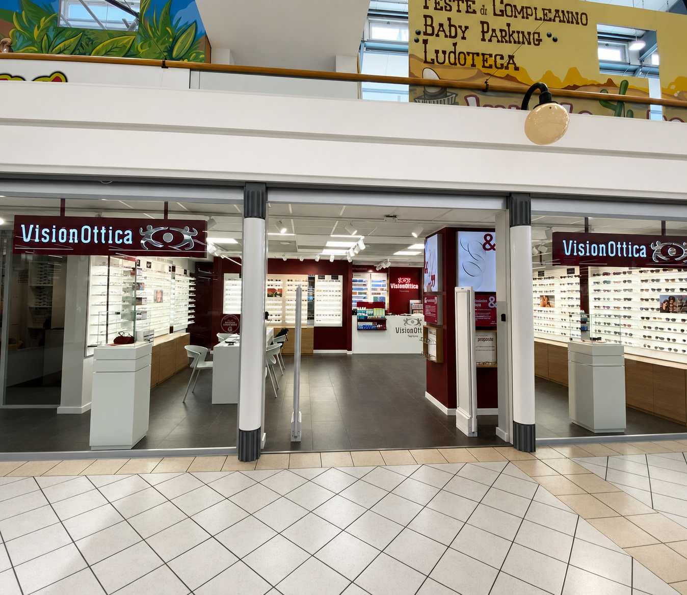 Ingresso del negozio VisionOttica Tiburtina di Roma in centro commerciale con ampia apertura su due livelli, insegna bordeaux bordeaux, espositori di occhiali su entrambe le pareti laterali e cartelli con temi per bambini sul soppalco