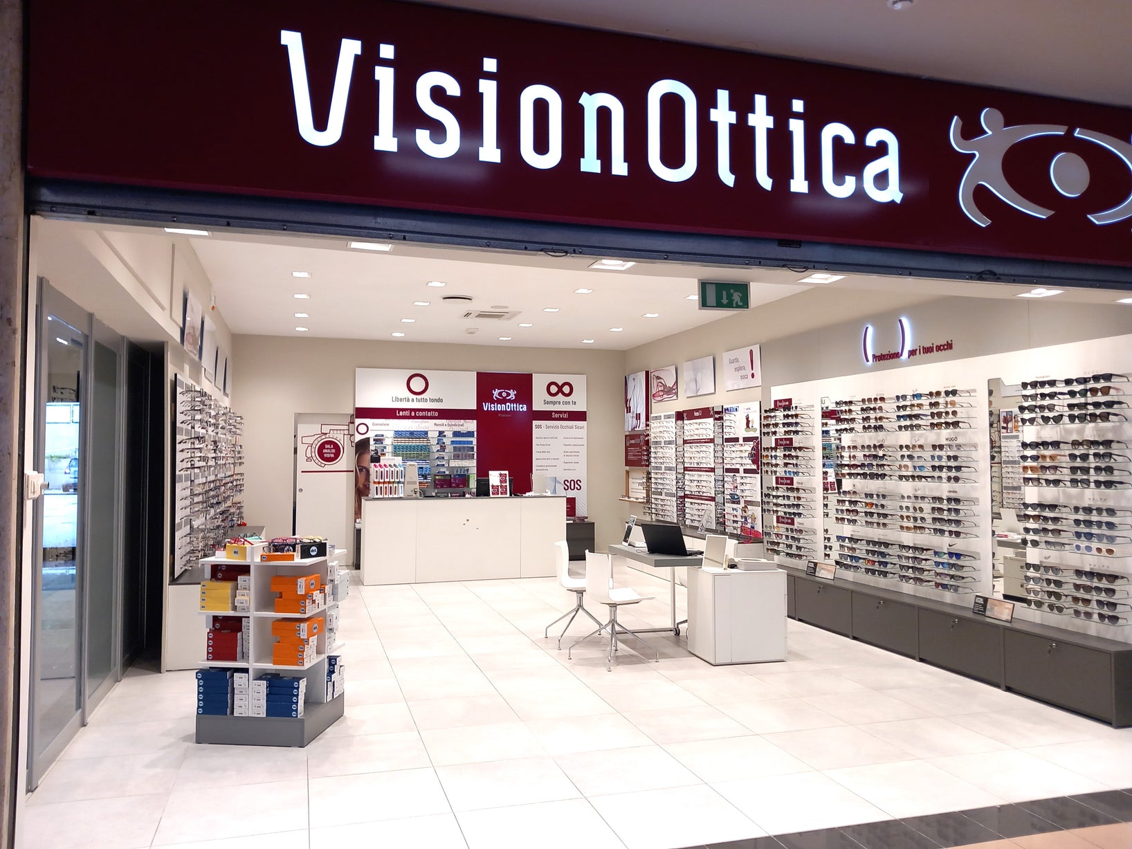 Ingresso del negozio VisionOttica Platani di Palestrina in centro commerciale con insegna luminosa bianca su fondo bordeaux, ampia apertura su interno con scaffali laterali di occhiali, area lenti a contatto e bancone bianco centrale