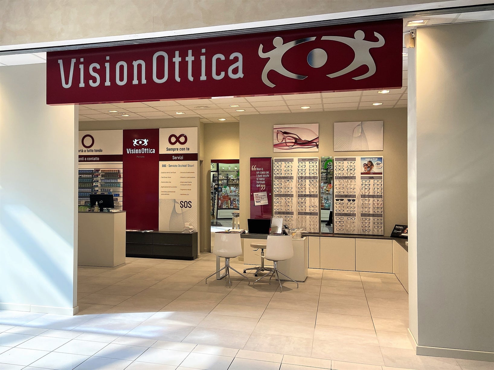 Ingresso del negozio VisionOttica Perseo di Rieti in centro commerciale con insegna bordeaux, apertura ampia su interno con scaffali di montature su parete bianca, bancone nero, area lenti a contatto e totem SOS centrale
