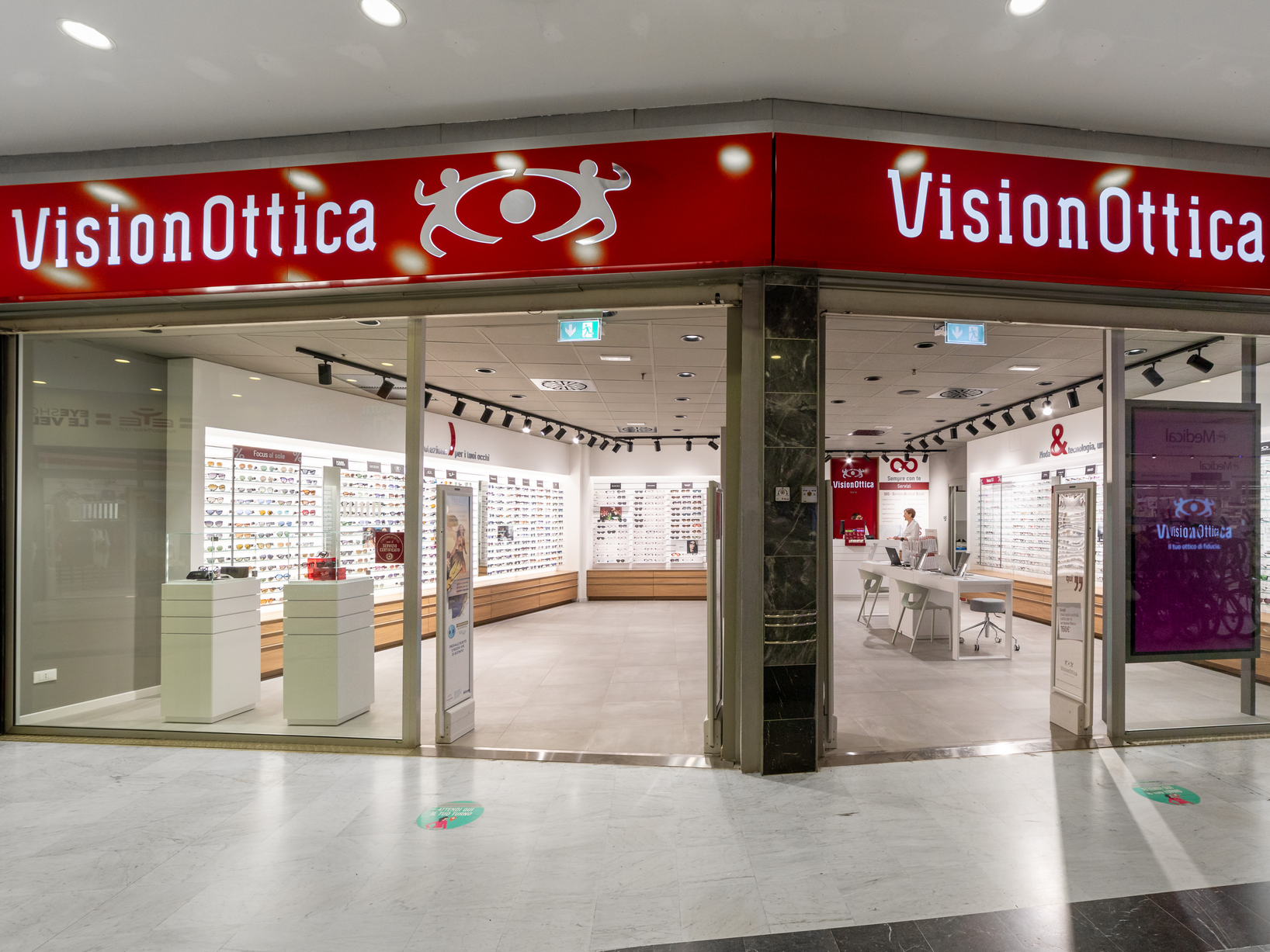 Facciata del negozio VisionOttica Le Vele di Quartu Sant'Elena in centro commerciale con doppia insegna rossa su ampia apertura a vetri, interno spazioso con espositori bianchi di occhiali su pareti, tavolo centrale e pannello viola con branding