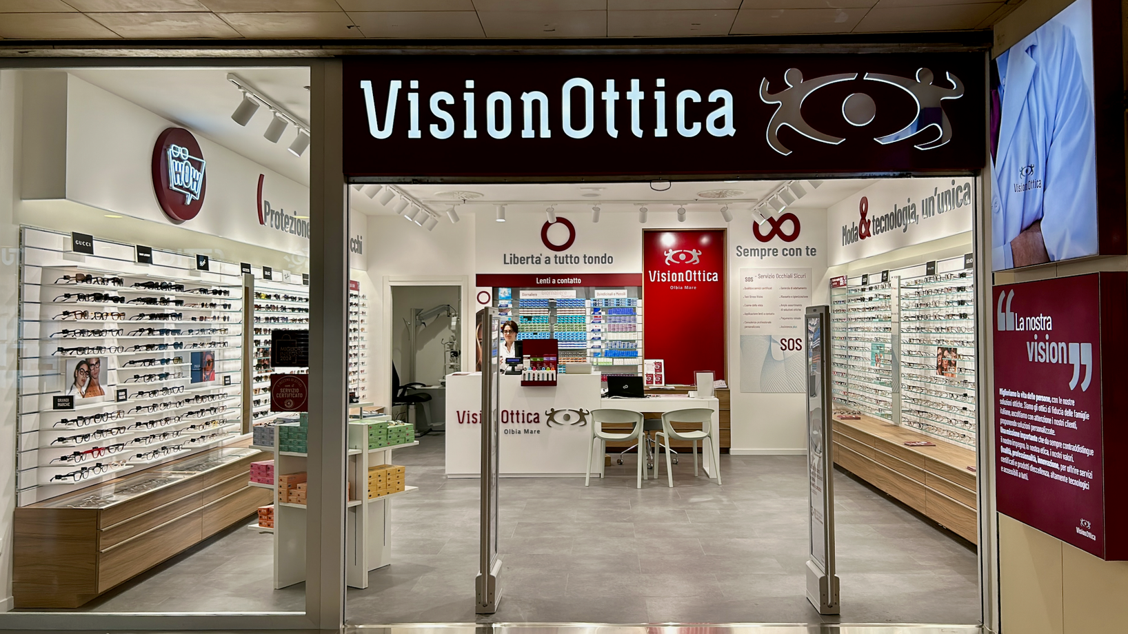 Ingresso del negozio VisionOttica Olbia Mare di Olbia in centro commerciale con insegna bianca su fondo bordeaux, ampia apertura su interno luminoso con scaffali laterali di occhiali, bancone centrale, pannello SOS e totem promozionale