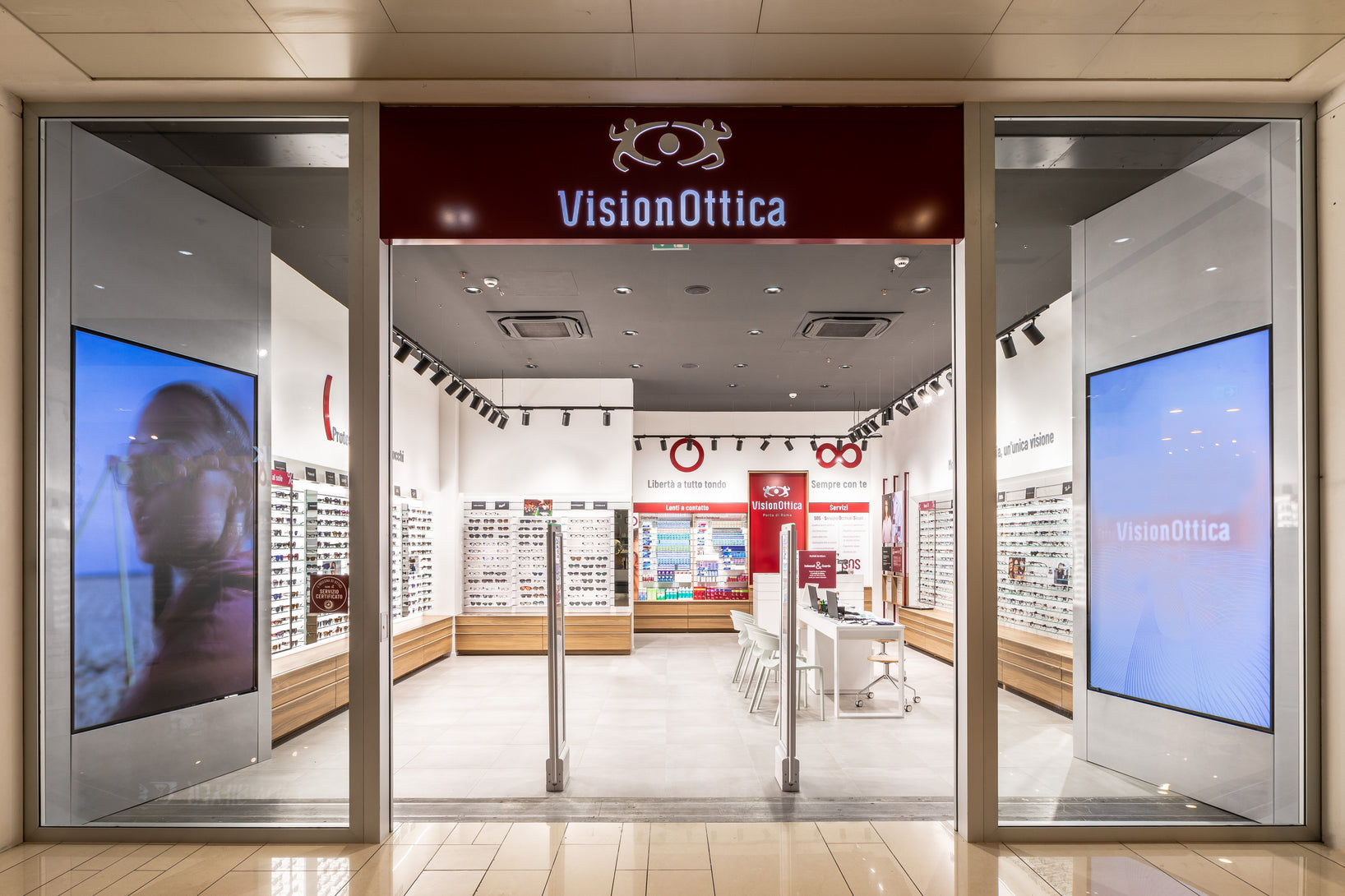 Ingresso del negozio VisionOttica Porta di Roma di Roma in centro commerciale con insegna bordeaux, ampia apertura con varchi antitaccheggio su interno con espositori in legno di montature, totem digitali laterali e bancone centrale rosso
