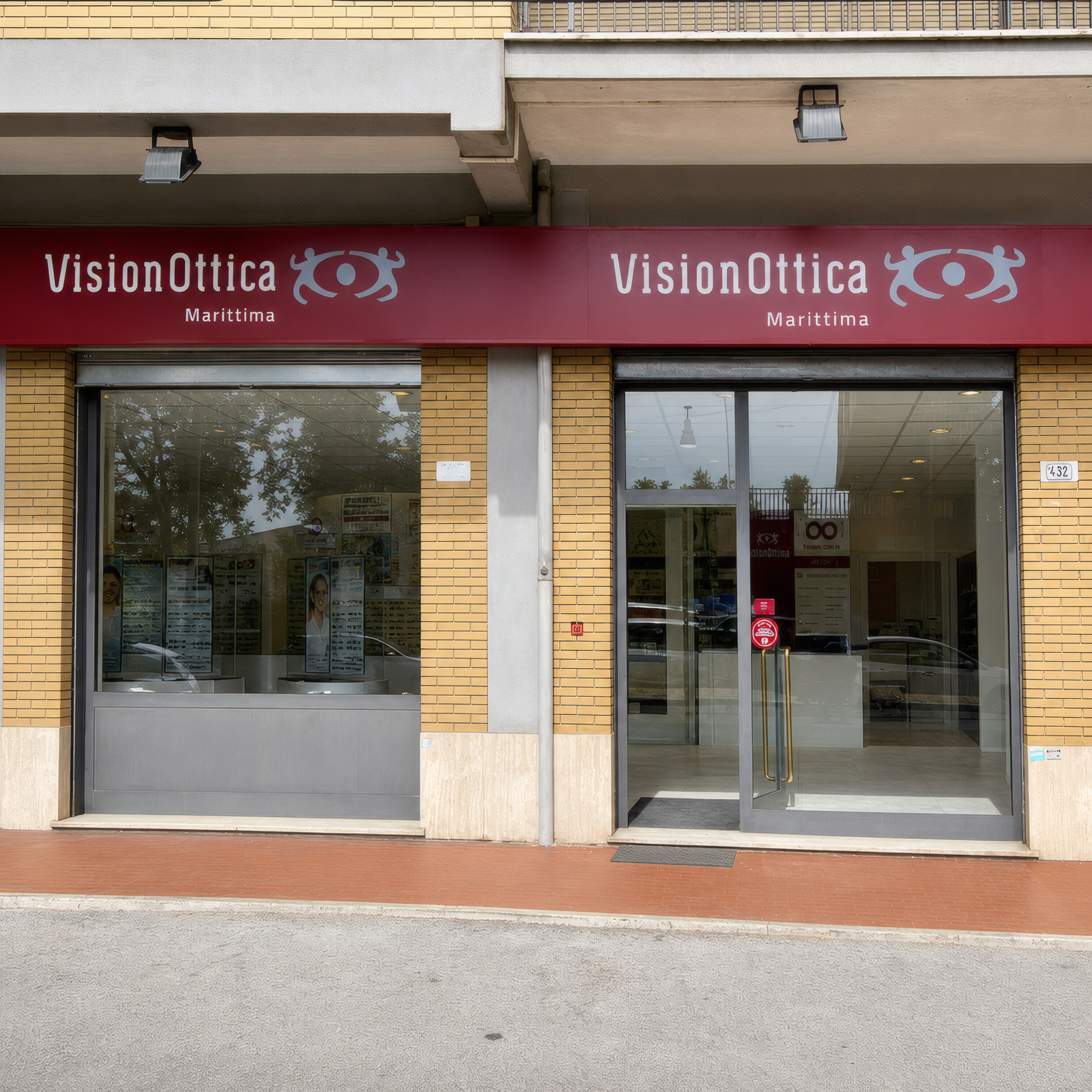Facciata del negozio VisionOttica Marittima di Frosinone su strada con doppia insegna bordeaux su edificio in mattoni gialli, due ingressi a vetri separati da pilastro e serrande abbassate lateralmente, marciapiede davanti