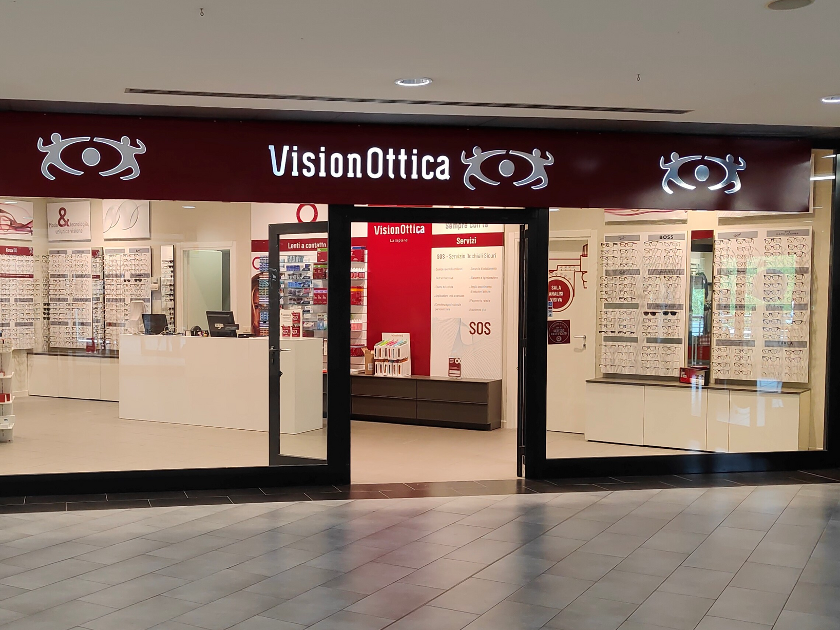 Ingresso del negozio VisionOttica Lampare di Arenzano in centro commerciale con insegna bordeaux orizzontale, porta a vetri centrale affiancata da espositori di montature su pareti bianche, bancone cassa e pannello servizi SOS visibili