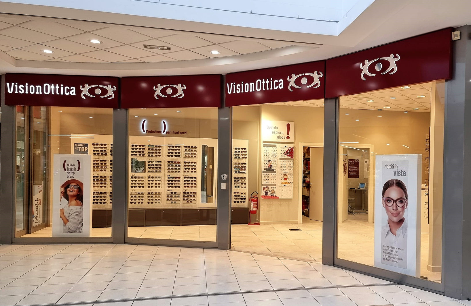 Facciata del negozio VisionOttica Mirto di Genova in centro commerciale con doppia insegna bordeaux su ampia apertura a due vetrate, interno con scaffali di montature su pareti, totem promozionali laterali con immagini di modelli e occhiali esposti