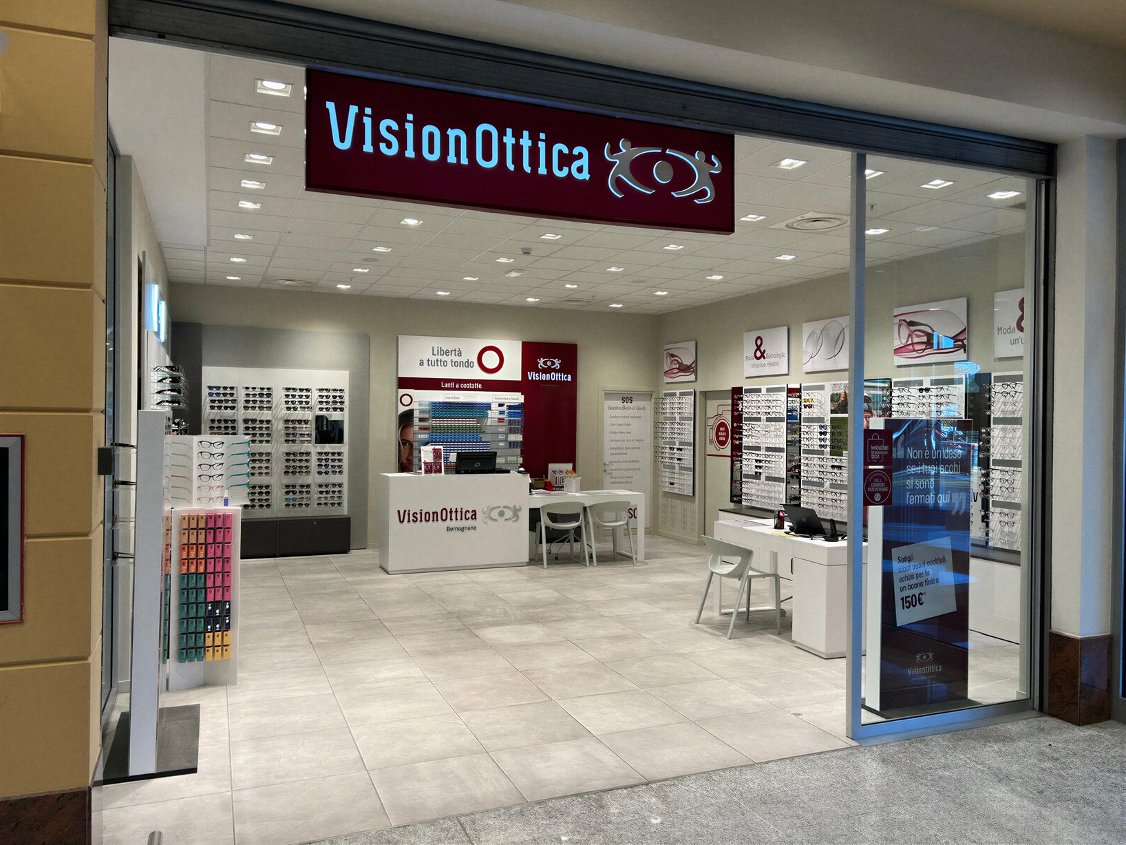 Ingresso del negozio VisionOttica Romagnano di Romagnano Sesia in centro commerciale con insegna blu, bancone centrale e lunghi espositori di montature sui lati